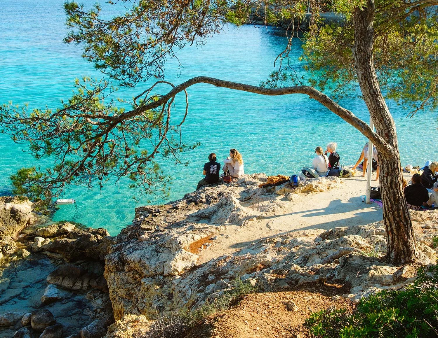 Menschen sitzen am Ufer eines klaren, blauen Sees, umgeben von Felsen und Bäumen, in einer sonnigen Küstenlandschaft.