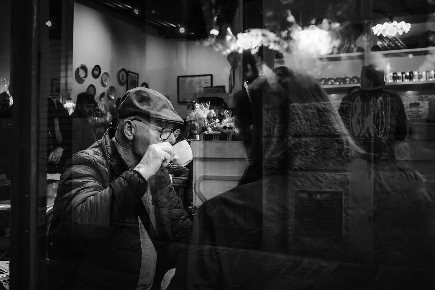Ein Mann mit Brille und Mütze trinkt in einem Café oder Restaurant, das durch eine Glasscheibe sichtbar ist. Die Szene ist schwarz-weiß, mit weiteren Personen im Hintergrund und Reflexionen auf dem Glas.