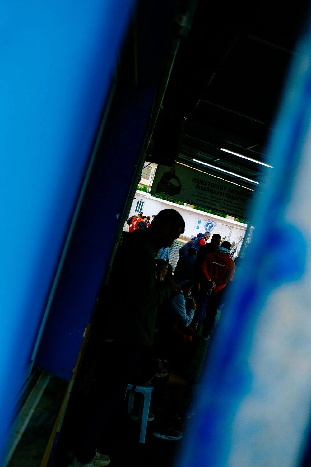 Menschen stehen und sitzen in einer Warteschlange im Freien, sichtbar durch einen schmalen Spalt. Im Hintergrund ist ein Schild mit griechischer Schrift zu erkennen.