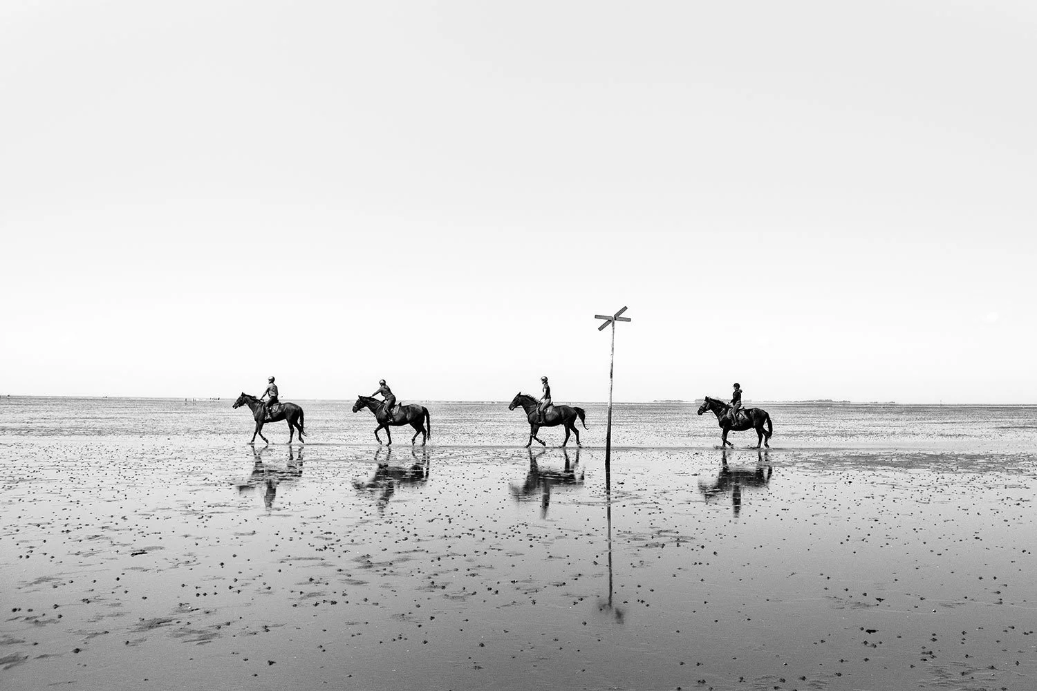 Vier Reiter auf Pferden reiten am Strand entlang, mit einem flachen Meer und einem leeren Himmel im Hintergrund, auf einem Strand mit reflektierenden Wasserstellen und einem einzigen Pfahl mit einerm Kreuz darauf.