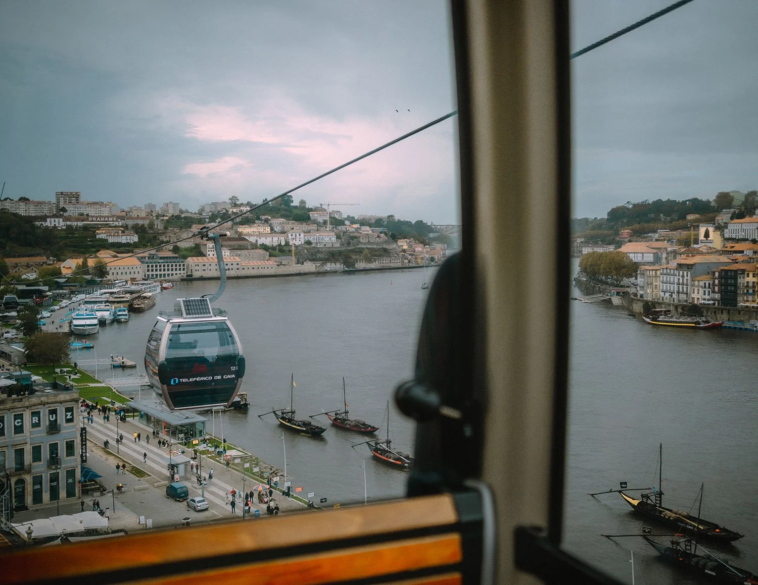 Blick aus einer Seilbahn auf eine Flussstadt mit Hängebrücke, Booten und einer Seilbahnstation am Ufer, bei bewölktem Himmel.