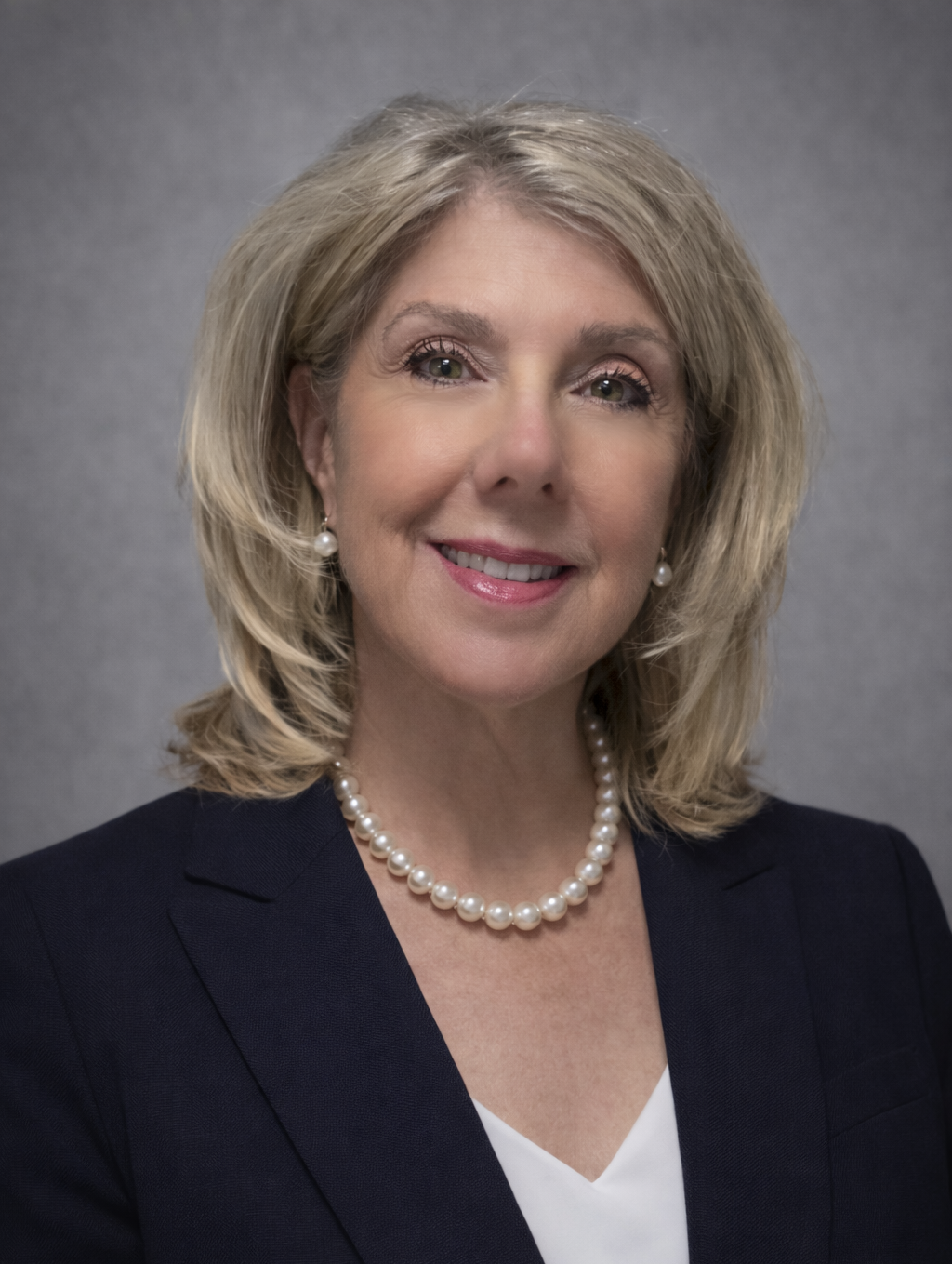 A portrait of a middle-aged woman with shoulder-length blonde hair, smiling, wearing a black blazer over a patterned top with a geometric design in black, white, blue, and beige, and a small necklace with a pendant.