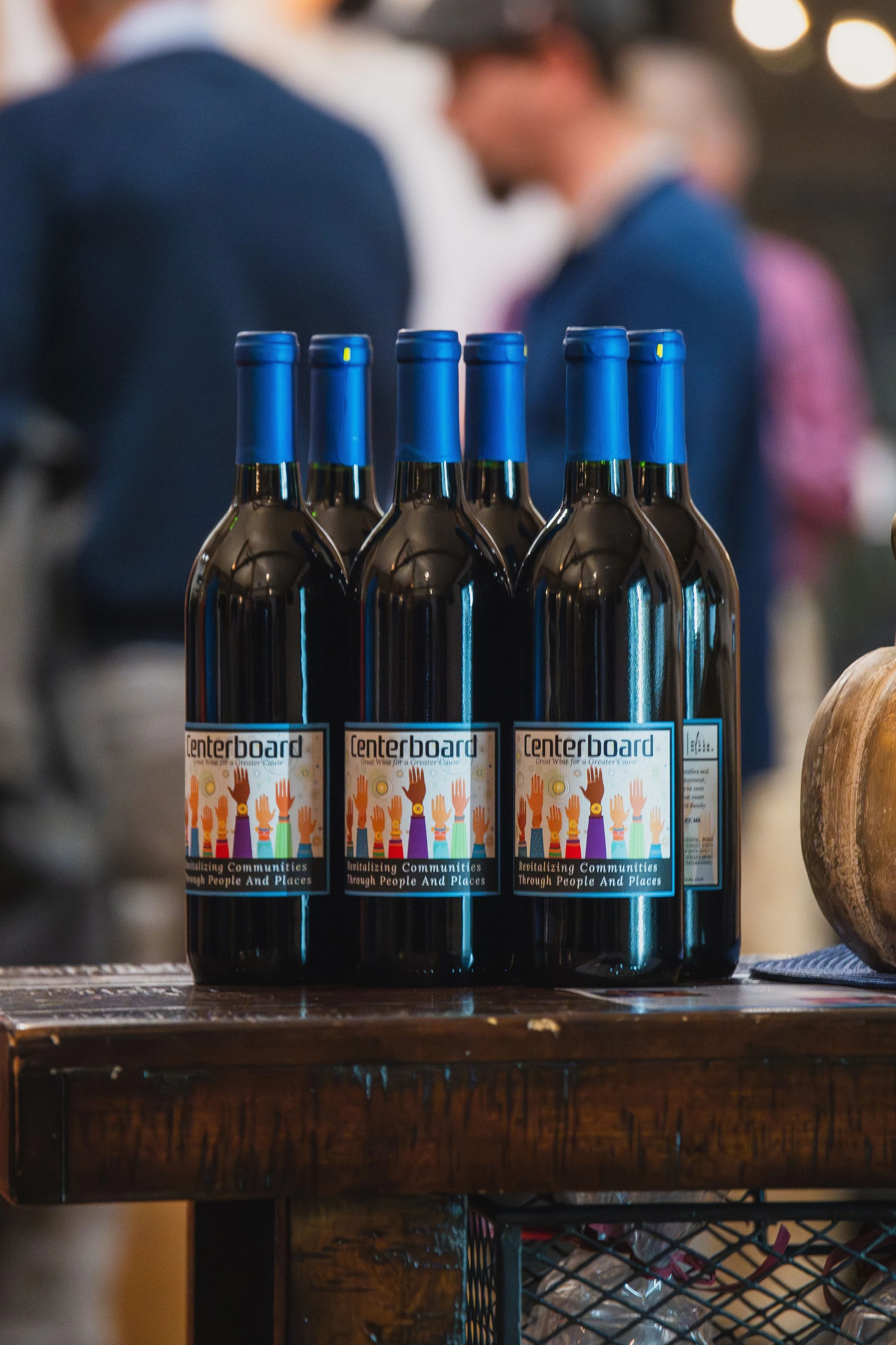 Four bottles of red wine with blue caps and labels displaying raised hands and text, placed on a wooden surface at an indoor event.
