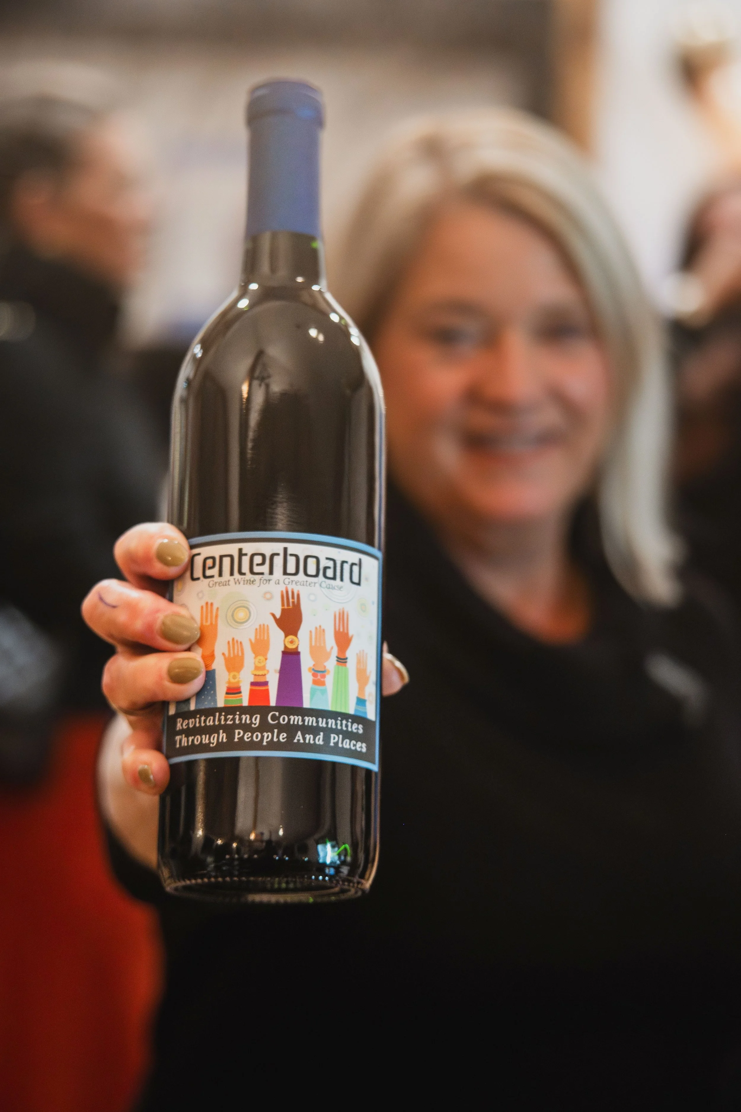 A woman with blonde hair holding a bottle of wine with a colorful label that reads "Penterboard- Great Wine for a Greater Cause." The label features illustrated raised hands of various colors and patterns. The woman is smiling and in the background, there are blurred people.