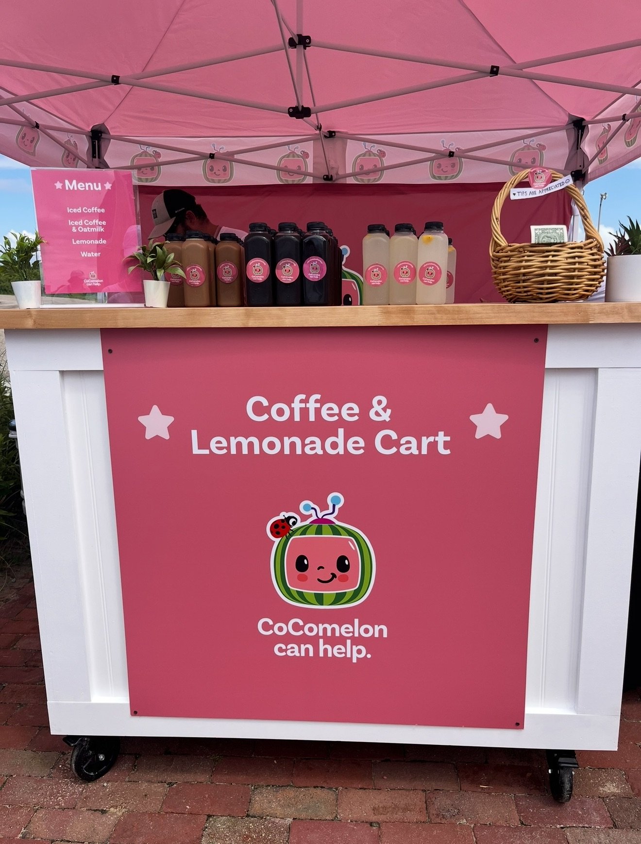 Two smiling people behind a pink and white cart with a sign that reads "Coffee & Lemonade Cart" and a cartoon watermelon logo. They are standing outdoors under a pink canopy, with one person wearing sunglasses and a white shirt, and the other person also wearing sunglasses and a white shirt. The cart has bottles of lemonade and coffee, with a basket and small potted plants on top.