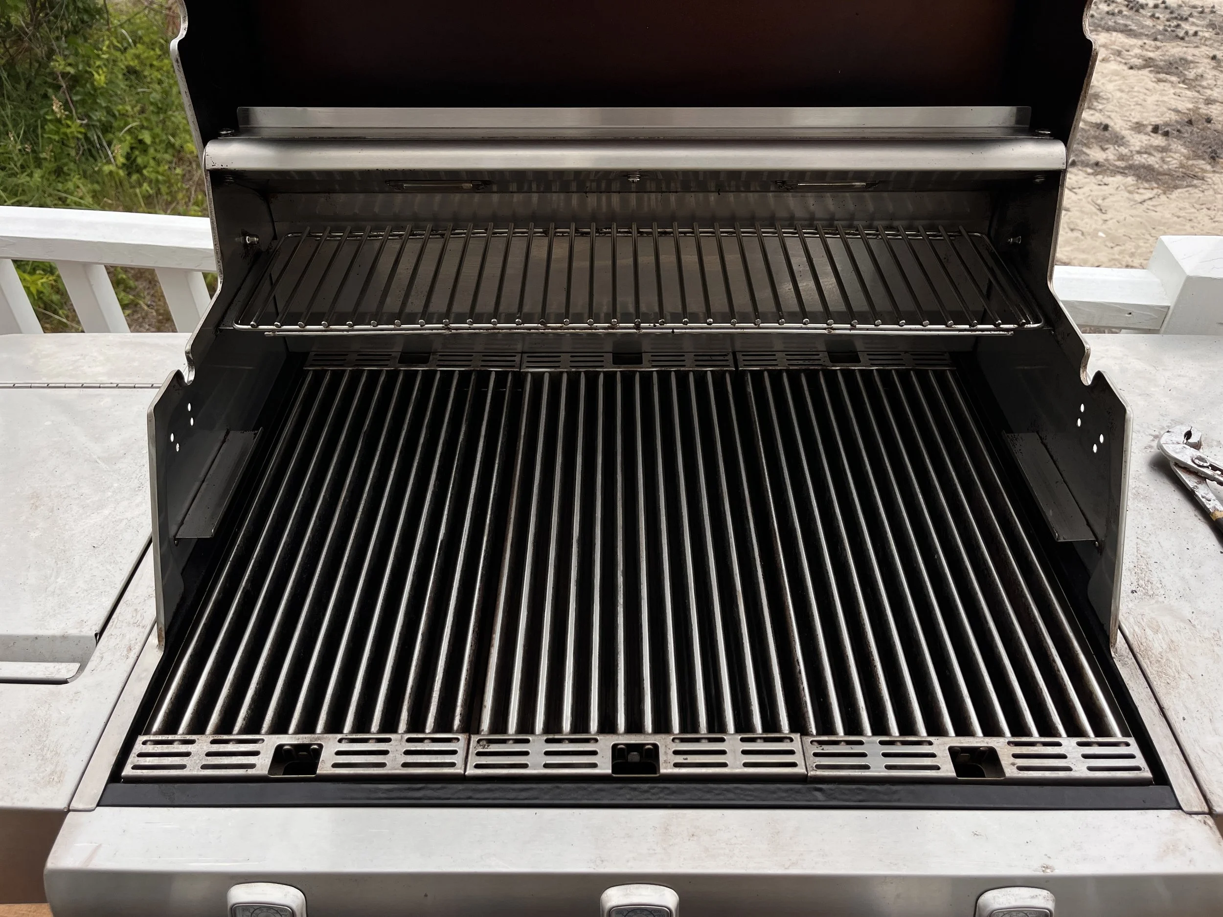 Open stainless steel gas grill with empty cooking grates, situated outdoors on a concrete surface.
