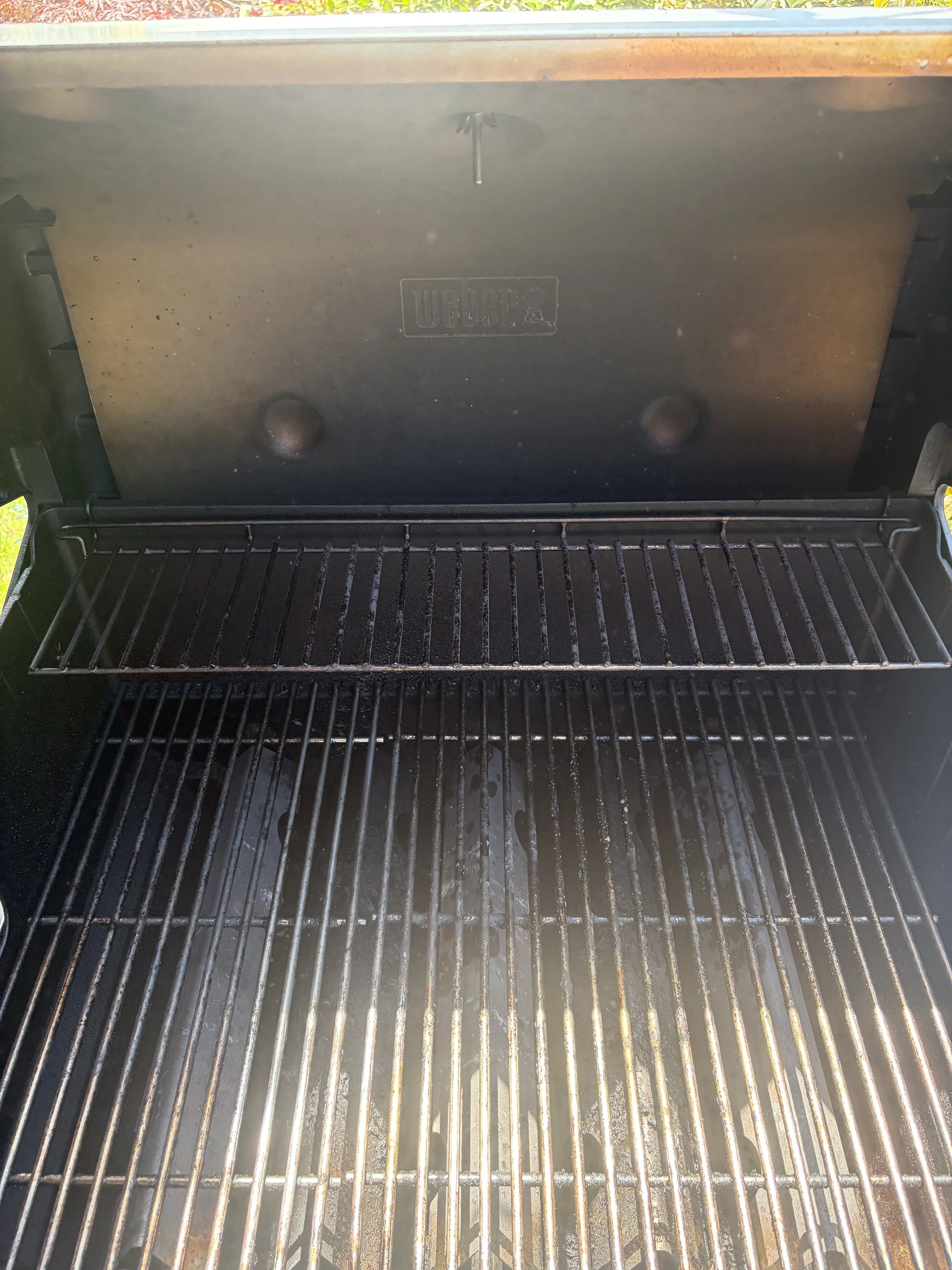 Close-up of a black grill inside an outdoor barbecue with a metal lid open. The grill grate has some rust and grease spots, and the lid shows the brand markings.
