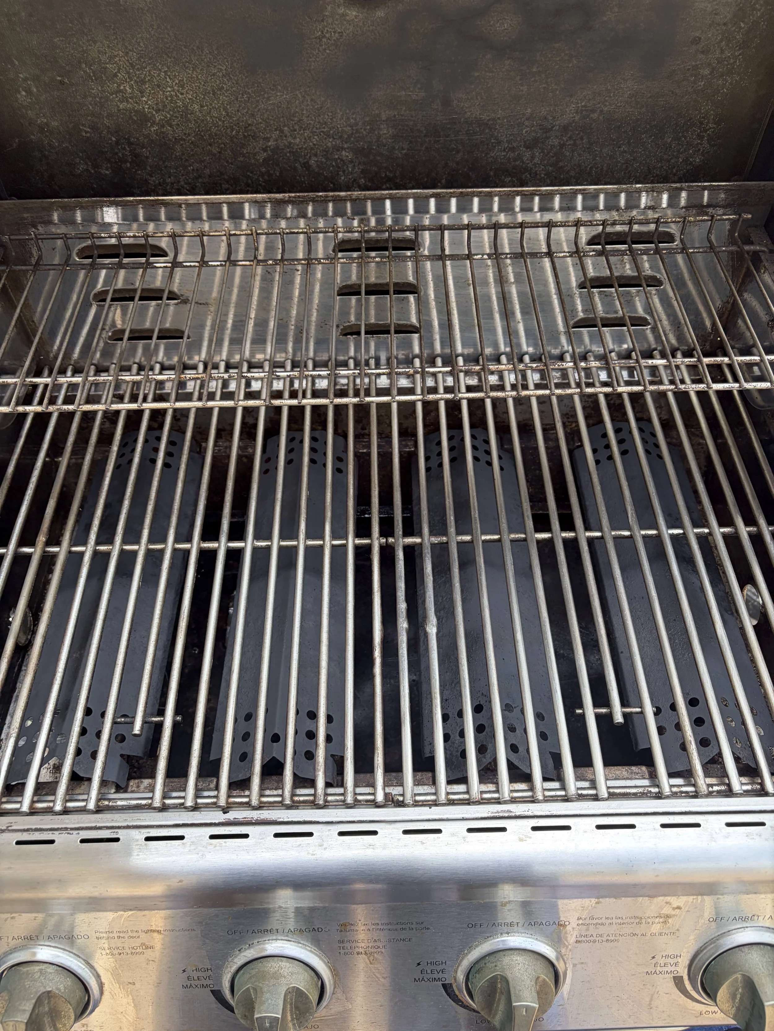 A stainless steel outdoor grill with four control knobs at the front and a metal grate on top. The knobs have labels in multiple languages and appear to be turned to different settings.
