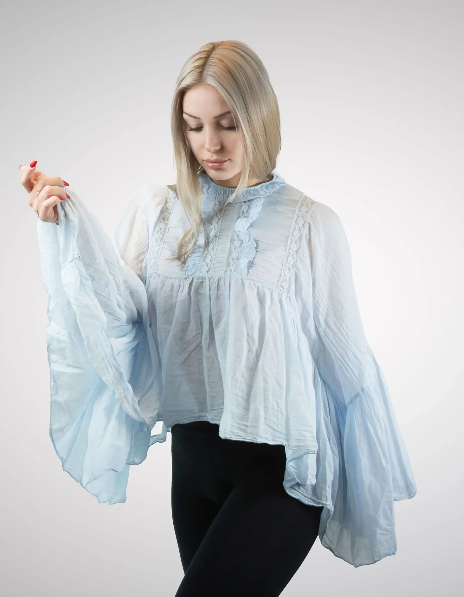 Modell trägt die voluminöse Boho-Bluse Clara in Hellblau mit filigranen Spitzeneinsätzen auf der Brust, hochgeschlossenem Kragen und weiten Glockenärmeln.