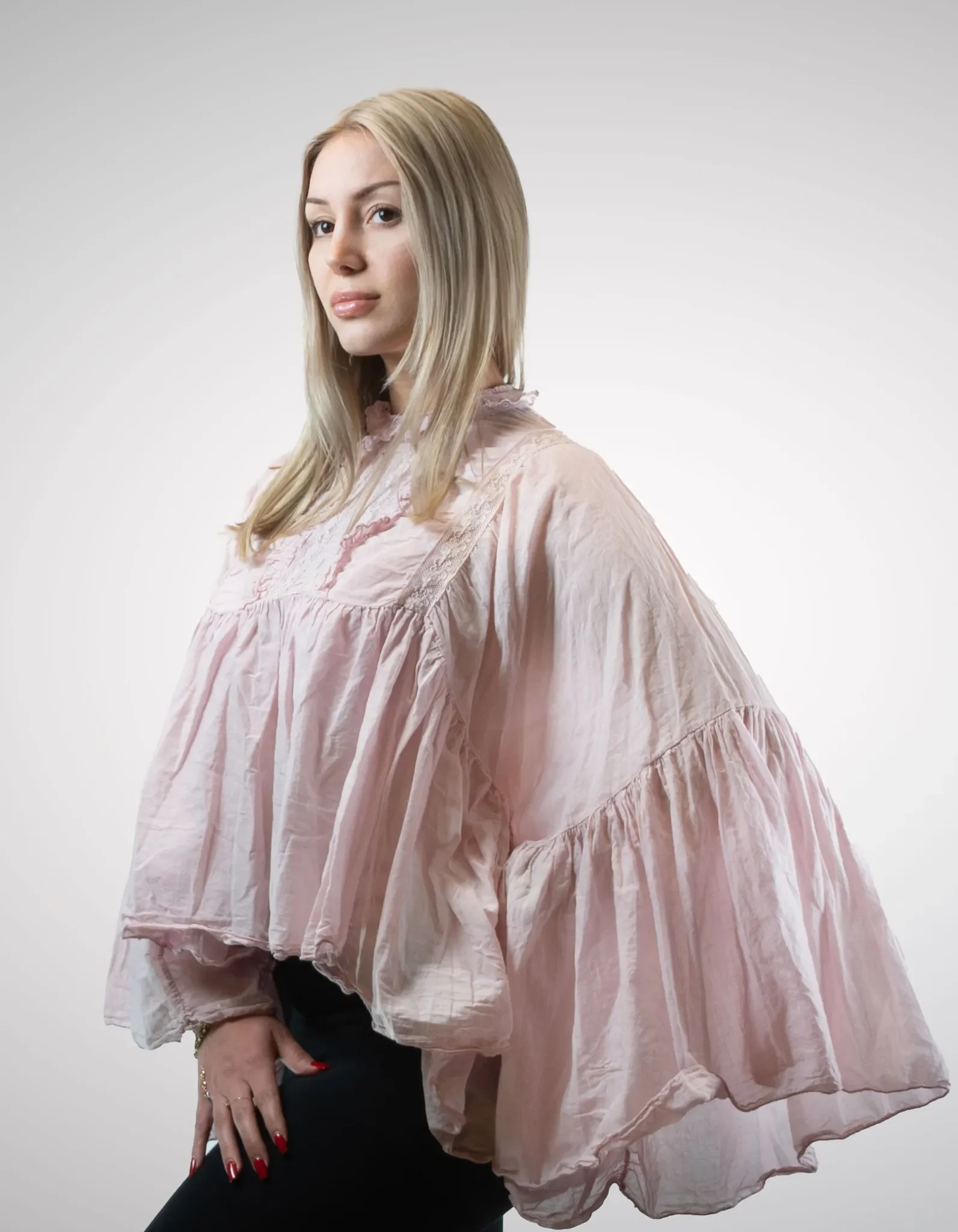 Modell trägt die voluminöse Boho-Bluse Clara in Zartrosa mit filigranen Spitzeneinsätzen auf der Brust, hochgeschlossenem Kragen und weiten Glockenärmeln.