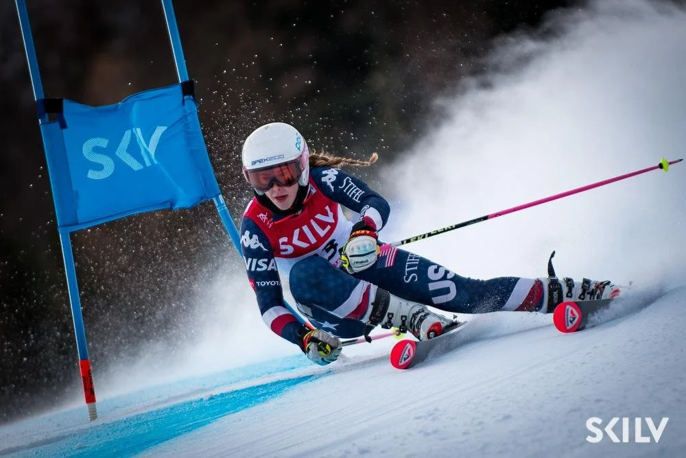A skier in blue gear, wearing a black helmet and reflective goggles, is racing down a snowy slope, making a sharp turn around a red gate during a ski competition.