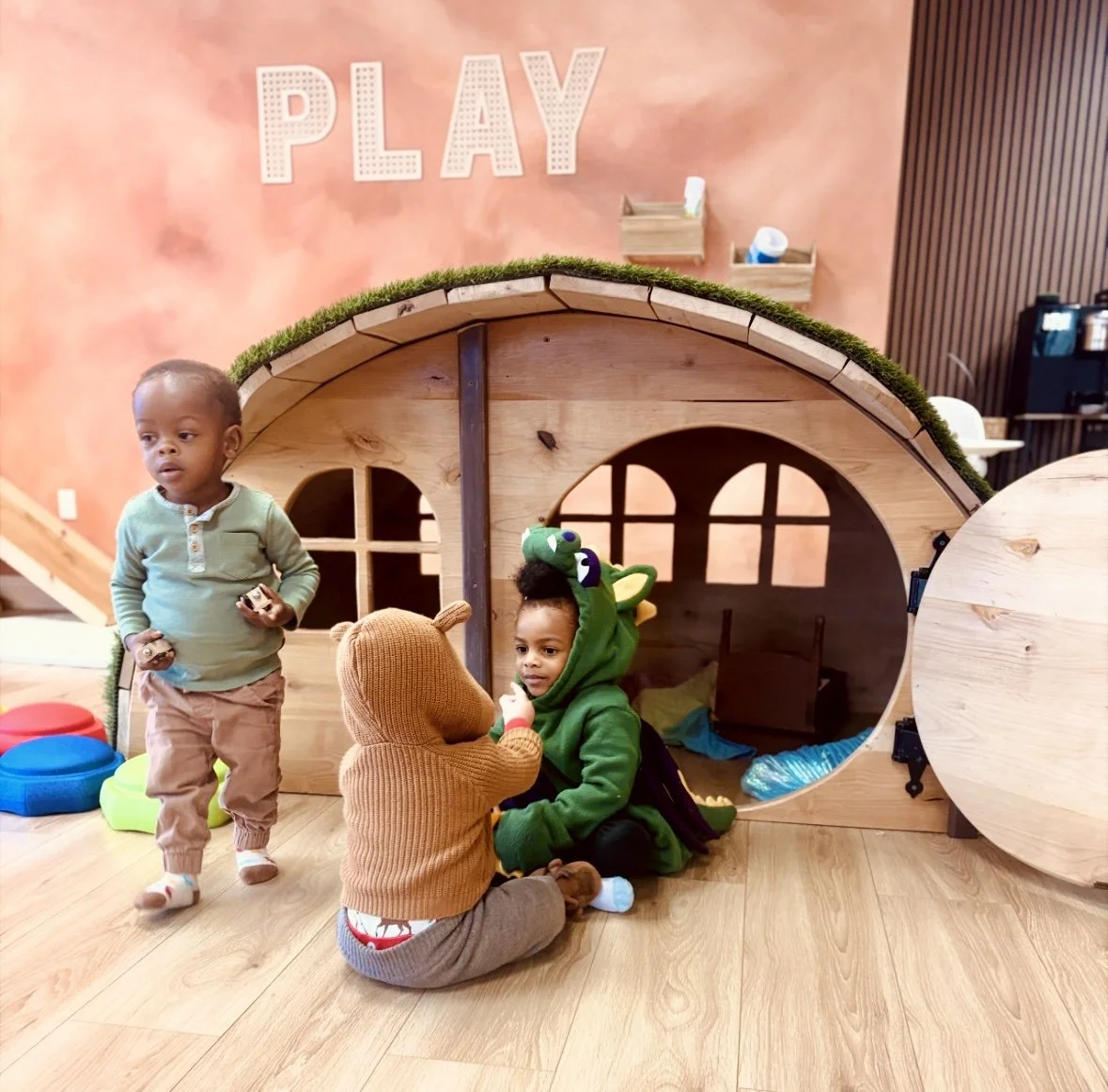 Kids enjoying open play at Kindred Play Collective, climbing in and around a wooden playhouse designed for imaginative toddler play.
