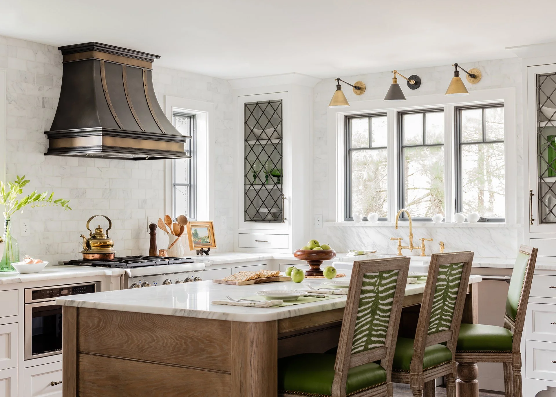 A bright kitchen with white marble countertops, a wooden kitchen island, and green upholstered chairs around a breakfast table. There are large windows, a brass faucet, a metallic range hood, and white cabinetry.