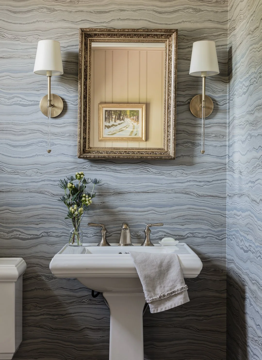 Dramatic powder room with marble swirl blue and grey wallcovering, pedestal sink, ornate gilt mirror, and brass sconces — luxury bathroom interior design in Essex County MA by Sacris Design