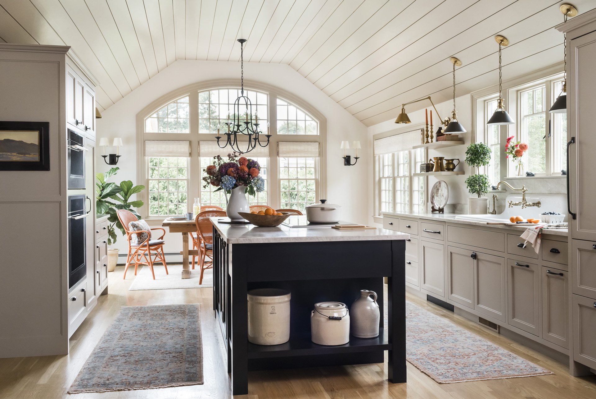 Beautiful kitchen with vaulted panelled ceiling, large arched window, brick house tan cabinets, black island with induction cooktop, vintage style sink with wall mounted faucet