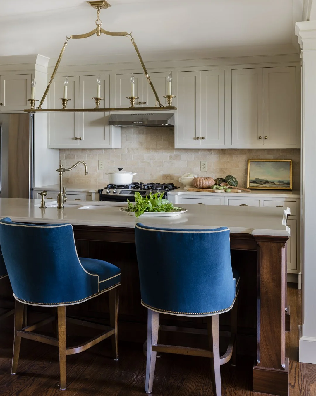 Luxury kitchen with mahogany island, peacock blue velvet barstools, cream shaker cabinetry, travertine stone backsplash, and brass candle chandelier — full service kitchen design in Essex County MA by SACRIS
