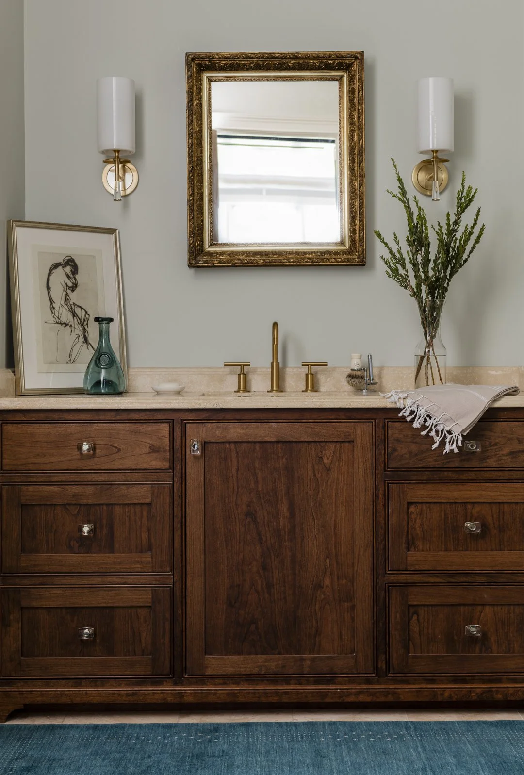 Primary bathroom vanity with dark wood cabinetry, travertine countertop, ornate gilt mirror, and brass sconces — luxury bath interior design in Essex County MA by Sacris Design