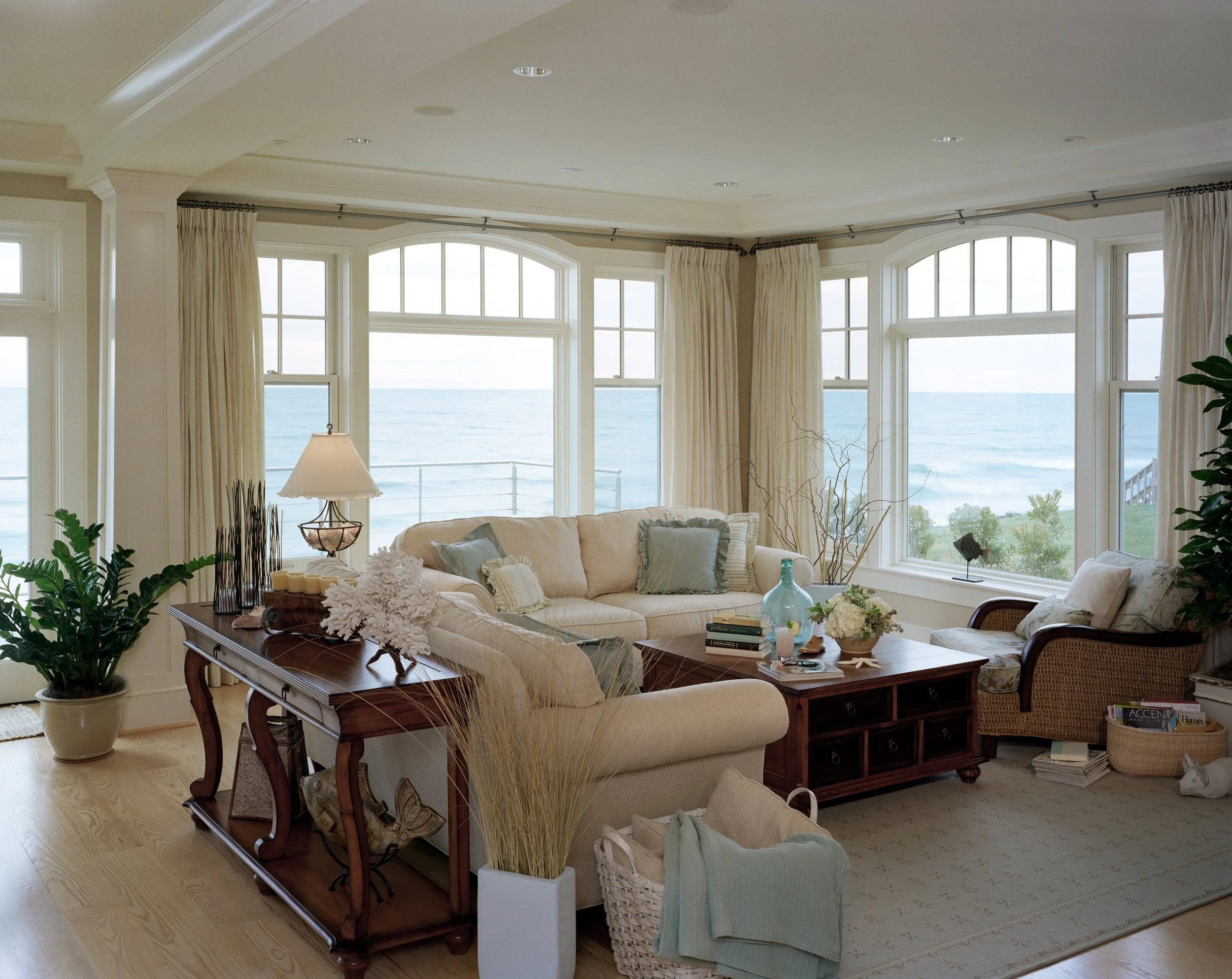 Coastal living room with panoramic ocean views, arched windows, cream upholstered seating, and antique mahogany console table — luxury beach house interior design in Rye, New Hampshire by SACRIS Interior Architecture & Design