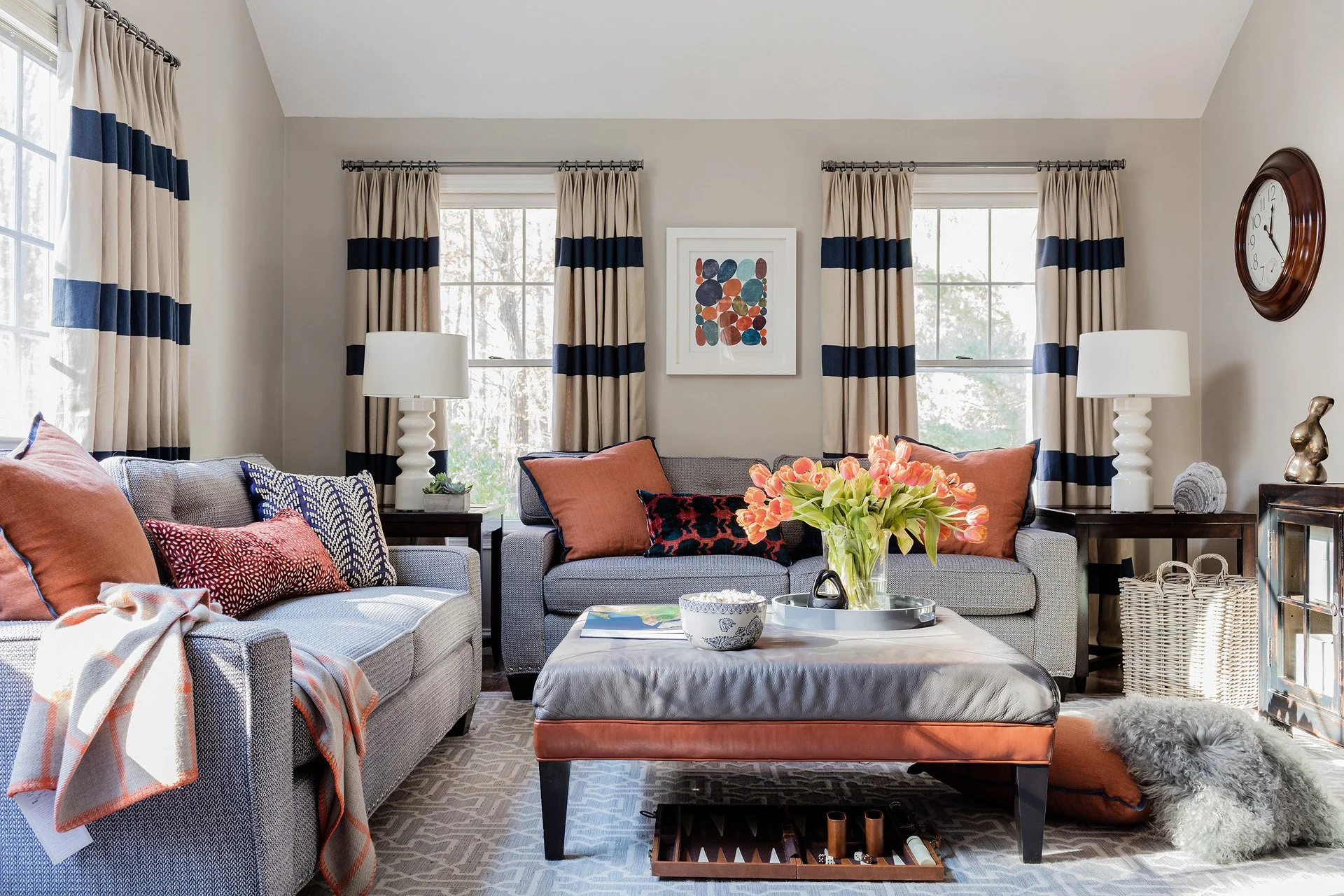 Living room with gray sofas, bold navy and cream stripe drapery, rust orange accent pillows, and leather ottoman — full service interior design in Boxford MA by Sacris Design