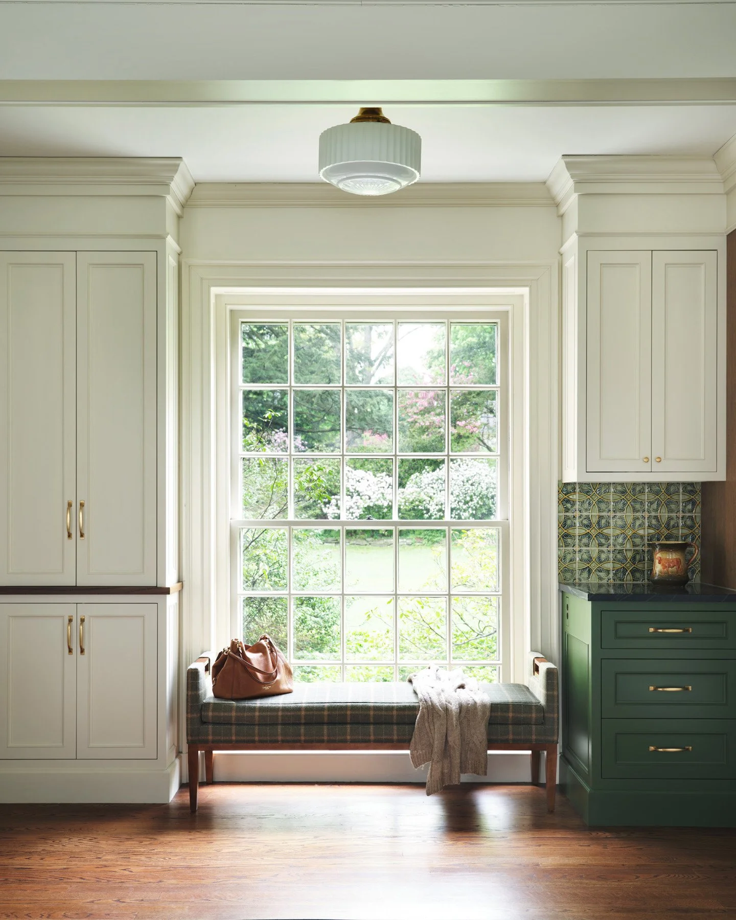 SACRIS kitchen window bench in plaid wool beneath an original multi-pane window, with cream inset cabinetry and vine-relief Arts and Crafts tile, Nashua New Hampshire