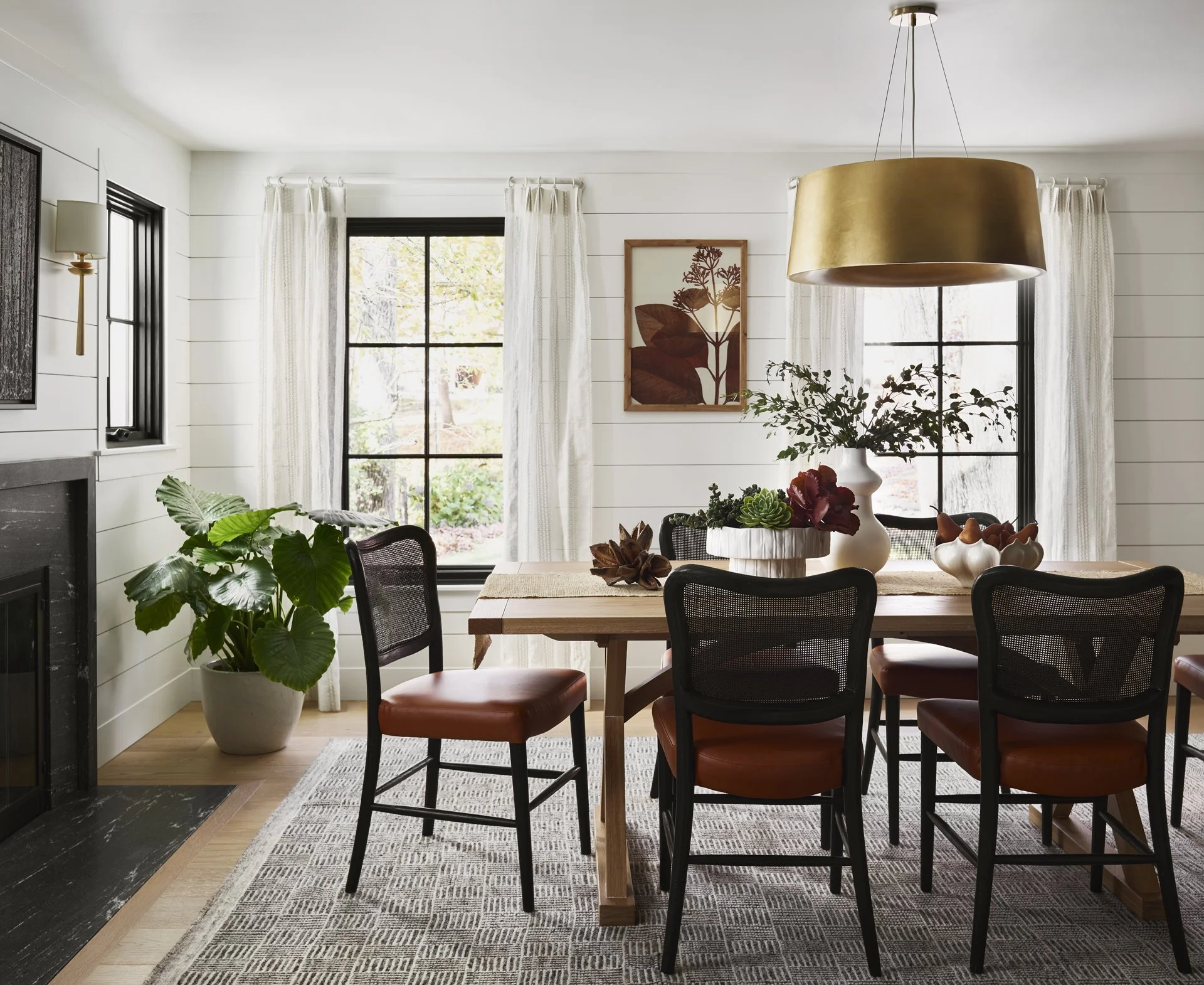 A dining room with a wooden oak table, black caned chairs with caramel leather cushions, shiplap wall panelling, black granite fireplace, visual comfort halo pendant chandelier