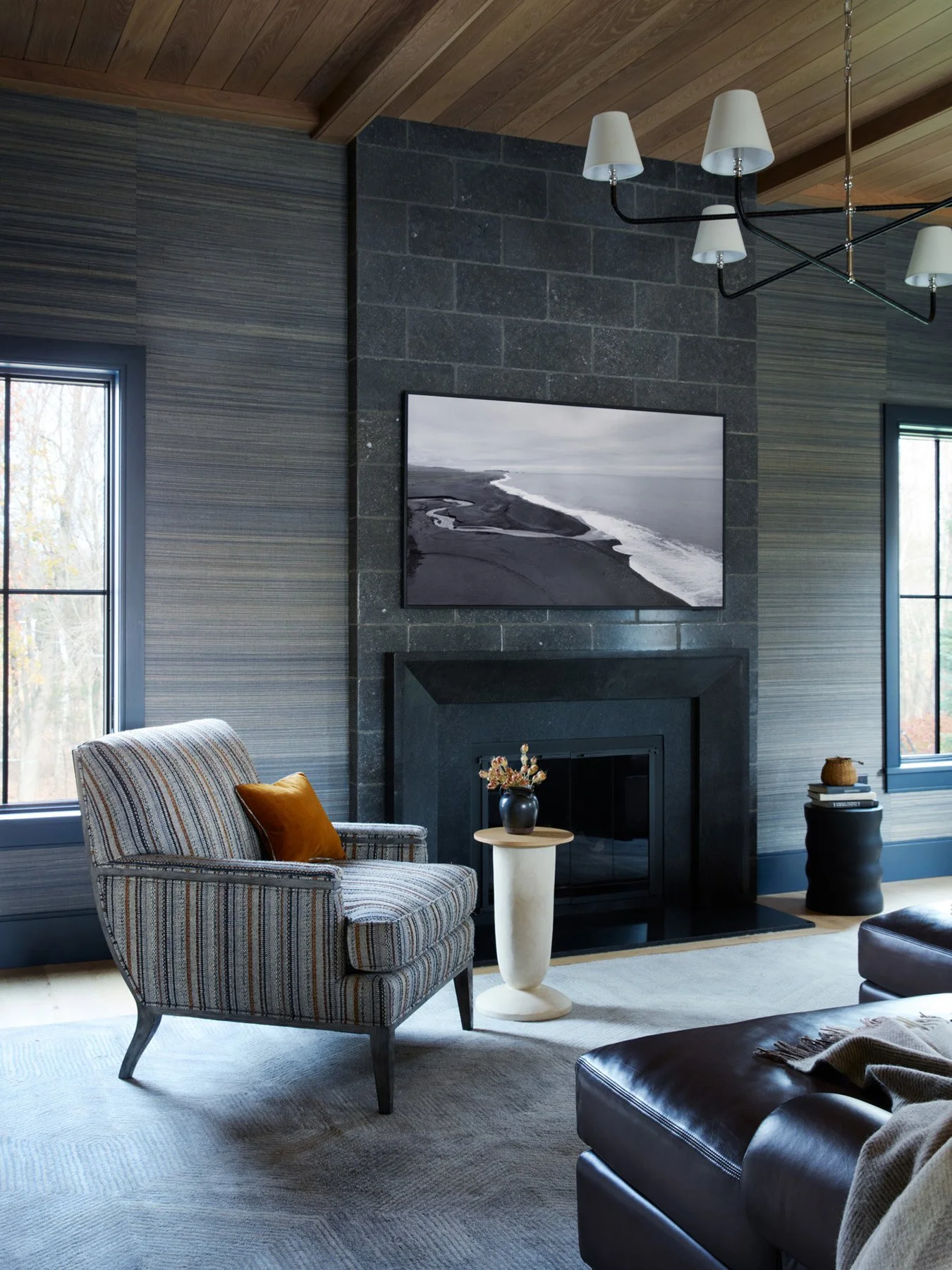 SACRIS great room fireplace in dark stone block with white oak ceiling, grasscloth walls, Visual Comfort Basden Grande chandelier and striped upholstered armchair, North Andover Massachusetts