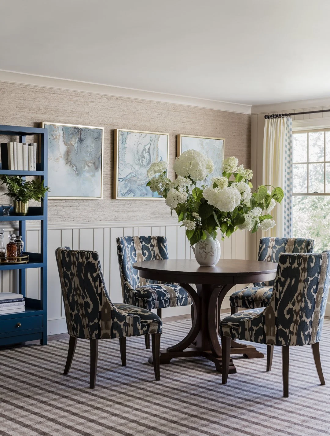 Traditional dining room with round dark pedestal table, blue ikat upholstered chairs, grasscloth wallcovering, and abstract art triptych — luxury interior design on the North Shore of Massachusetts by Sacris Design