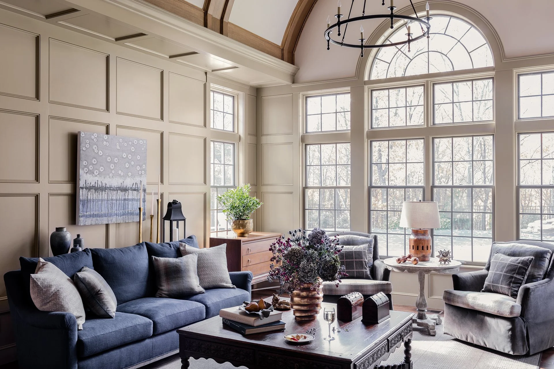 Living room with large arched windows, beige paneled walls, dark blue sofa with gray and beige pillows, coffee table with books, a flower arrangement, candles, and decorative objects, and a chandelier hanging from the ceiling