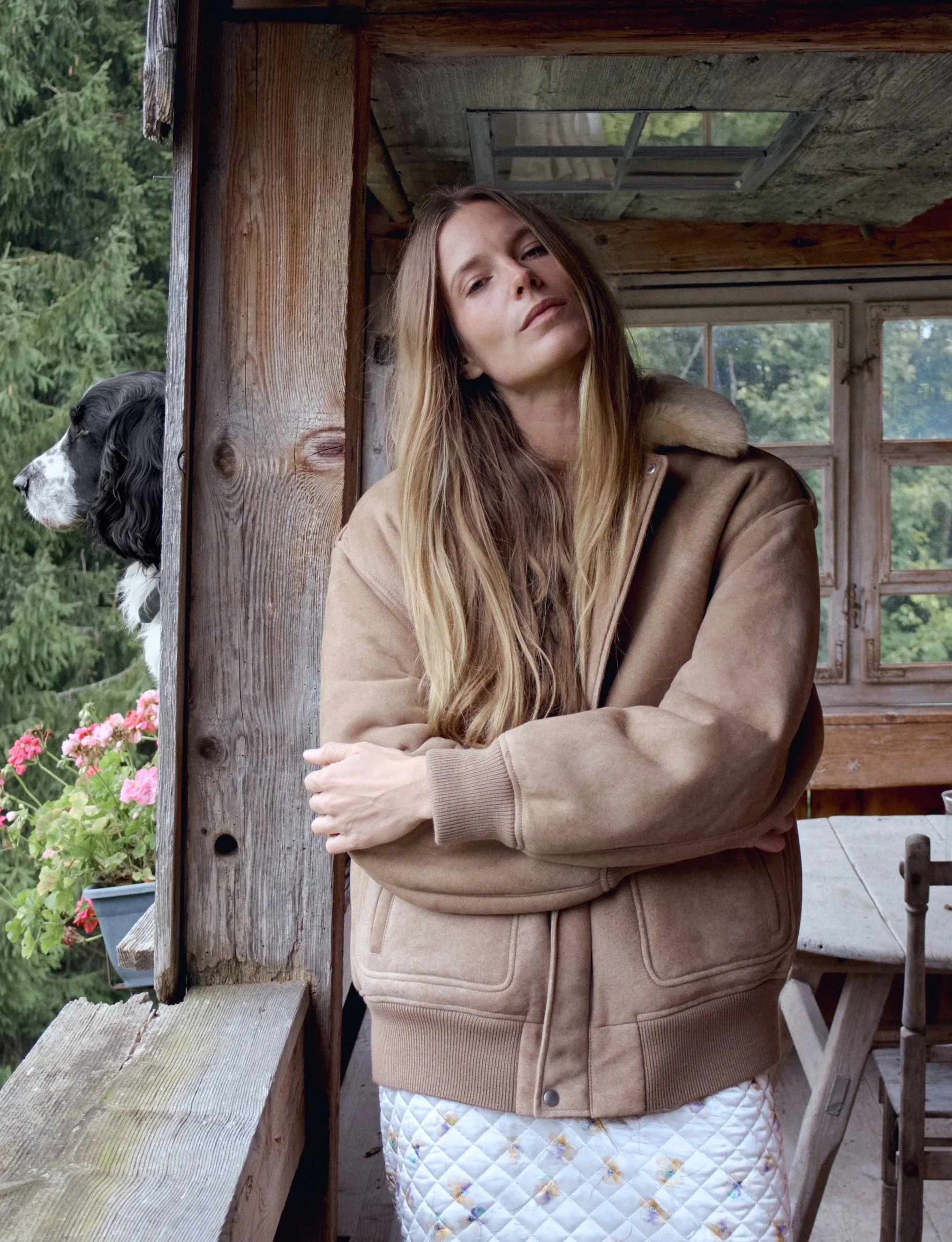 A woman with long hair wearing a tan jacket and a quilted skirt stands with arms crossed inside a wooden structure, with a dog peeking from behind a wooden post and greenery visible outside.