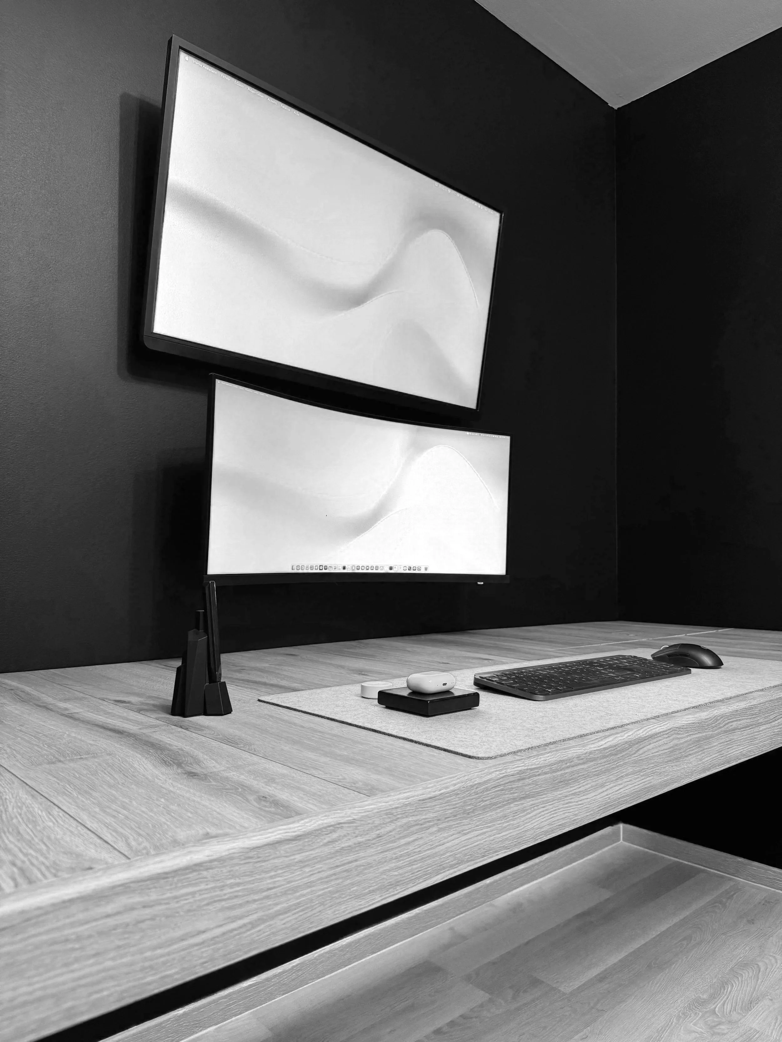 Un bureau avec deux écrans d'ordinateur montés sur un mur noir, un clavier, une souris, une télécommande, et des écouteurs sans fil placés sur la surface en bois clair.