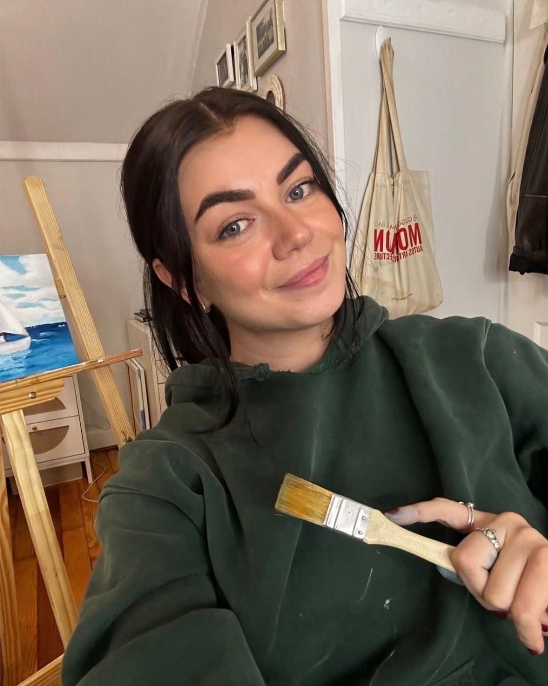 A woman with dark hair and blue eyes taking a selfie in a room with paintings, a bag on the wall, and an easel with a painting of a sailboat on water. She is holding a paintbrush in her hand and wearing a dark green hoodie.