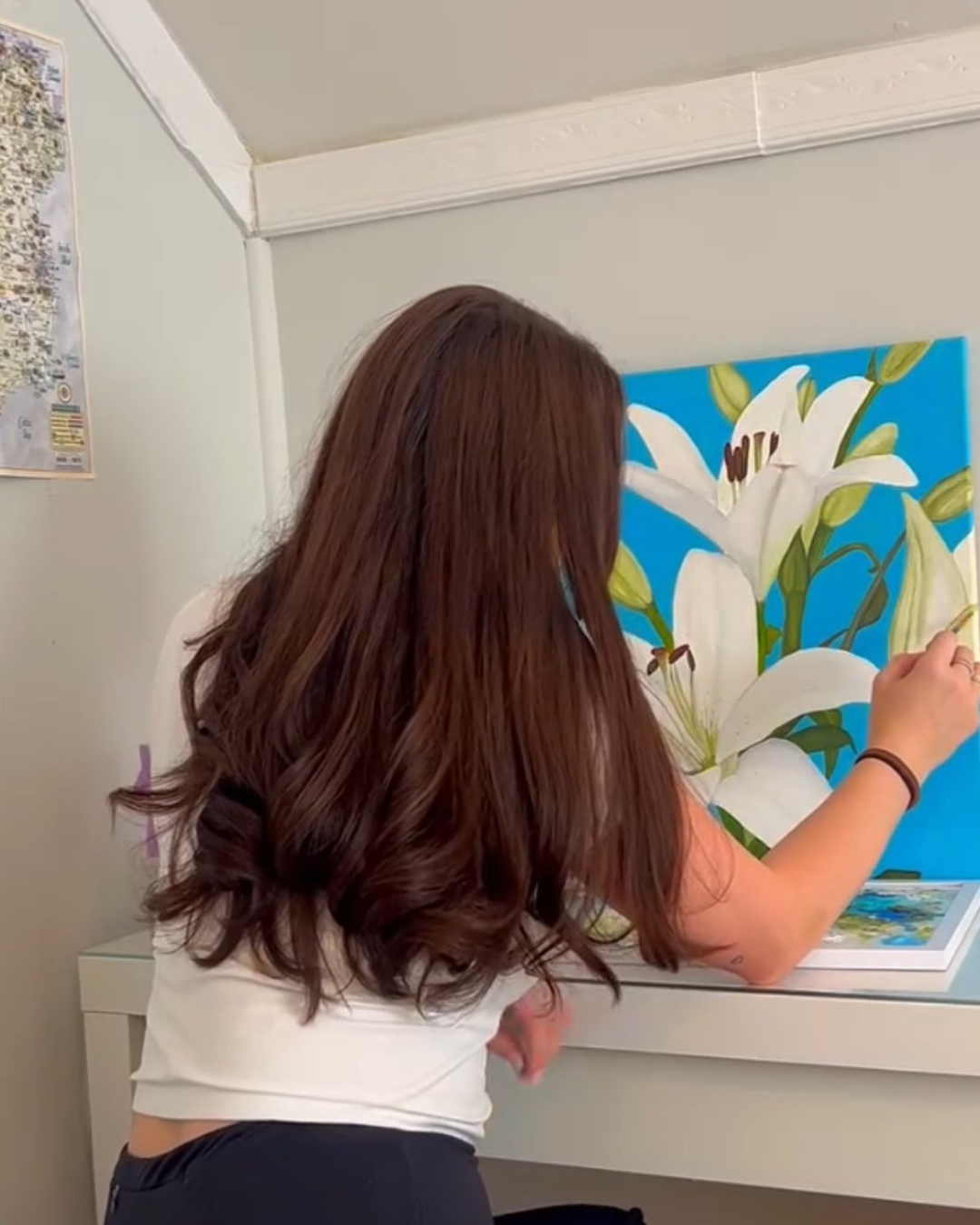 A woman with long, wavy, auburn hair painting a large canvas of white lilies with green leaves and a bright blue background. She is leaning over a white table.