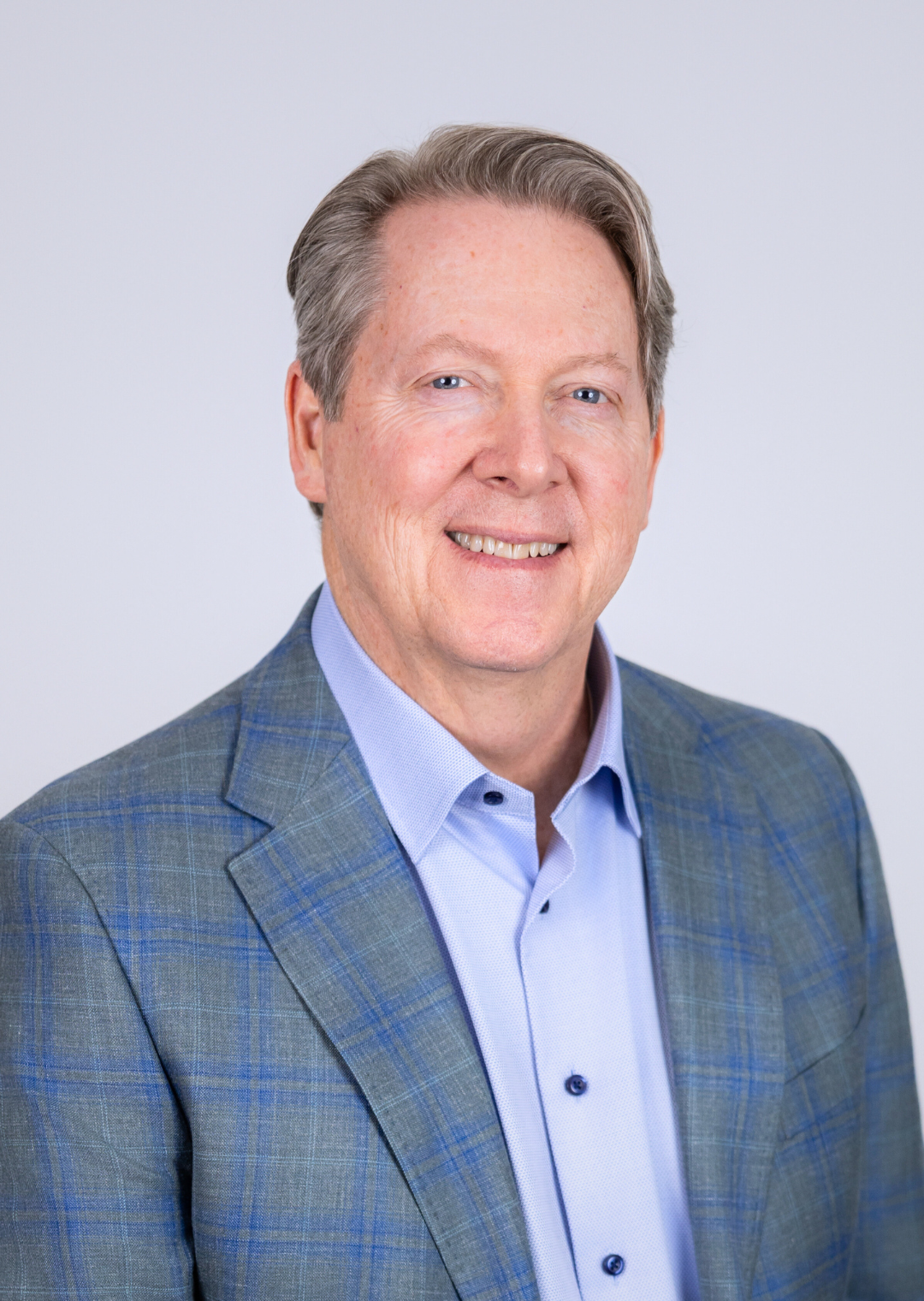 A professional headshot of Roger Pricer. He is wearing a blue shirt with a blue and grey tartan suitcoat