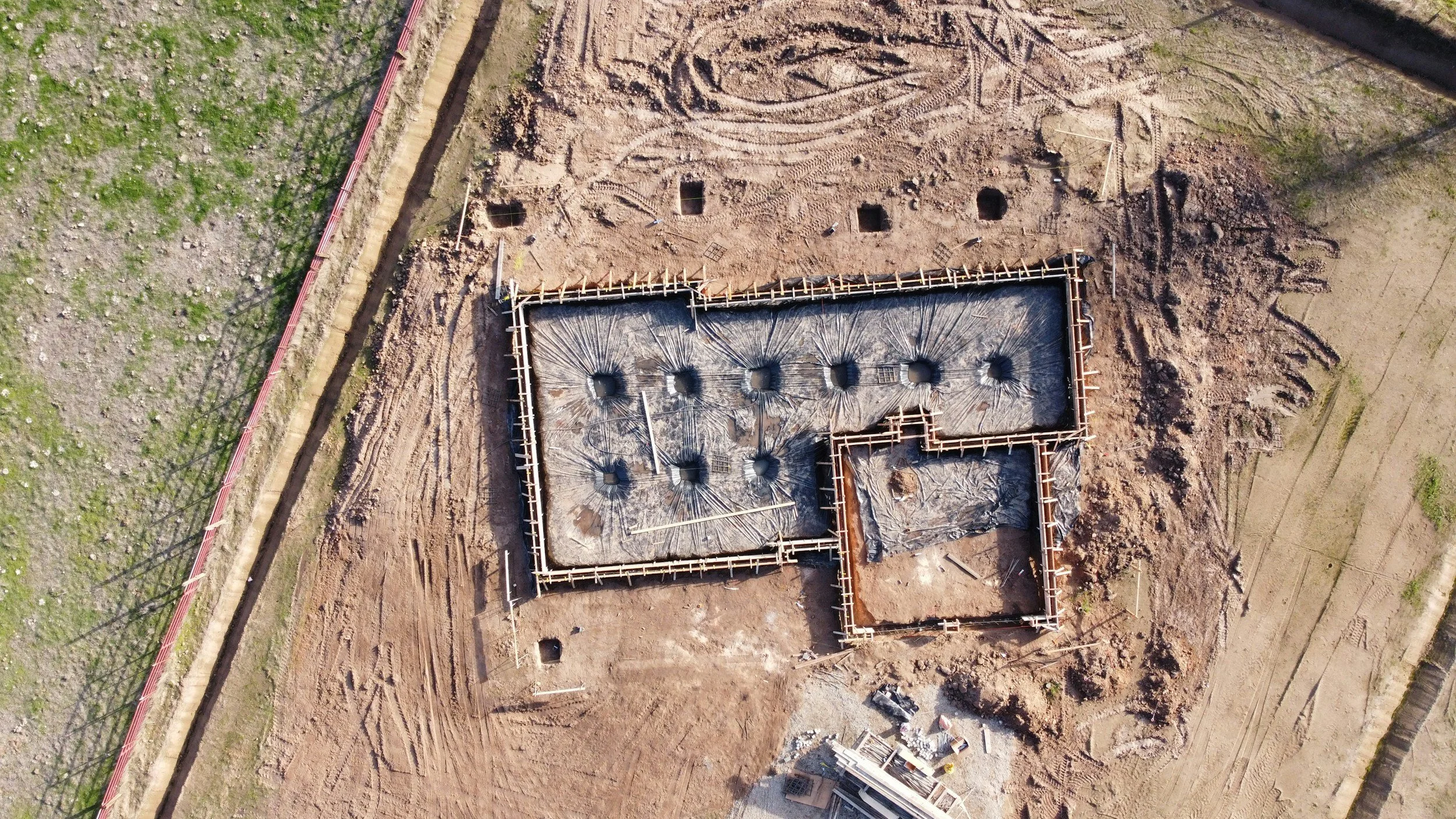 Aerial view of a construction site with a foundation outline marked on the ground, partially excavated earth, and construction materials scattered around.