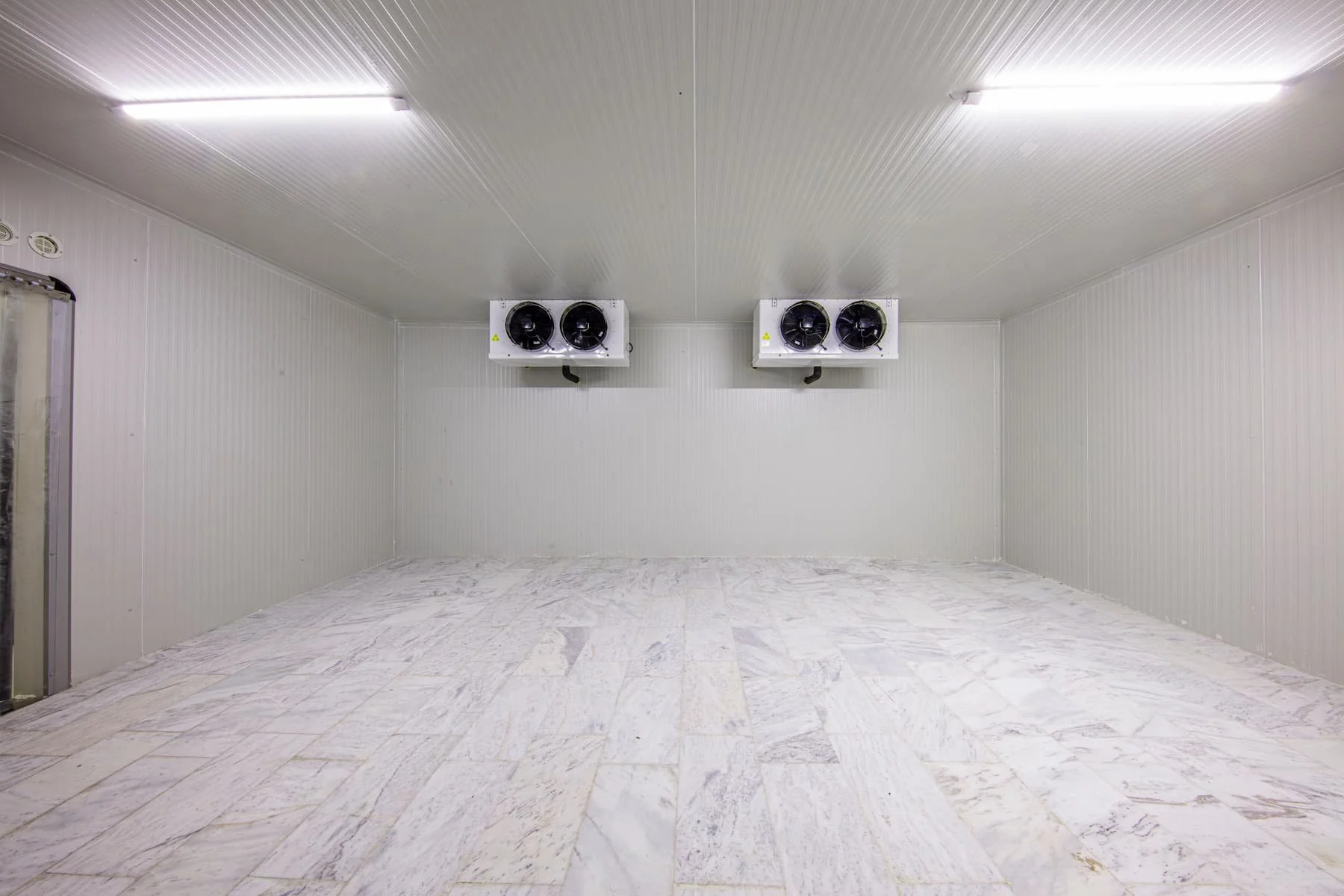 Empty commercial walk-in freezer with white walls, tiled floor, and overhead lighting. Two industrial cooling units mounted on the back wall.