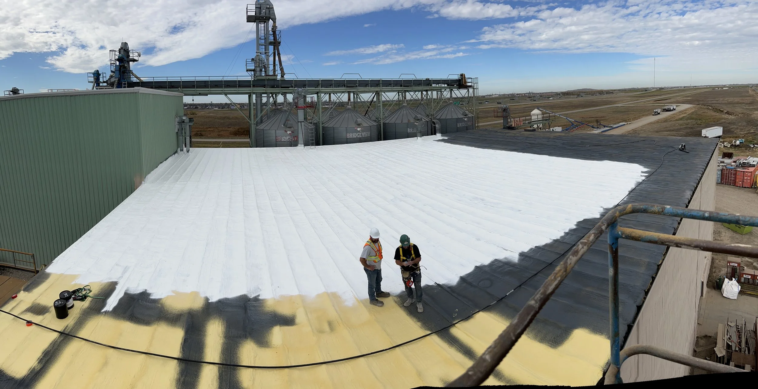 Two workers on a flat spray foamed roof with white silicone, with industrial equipment and silos in the background.