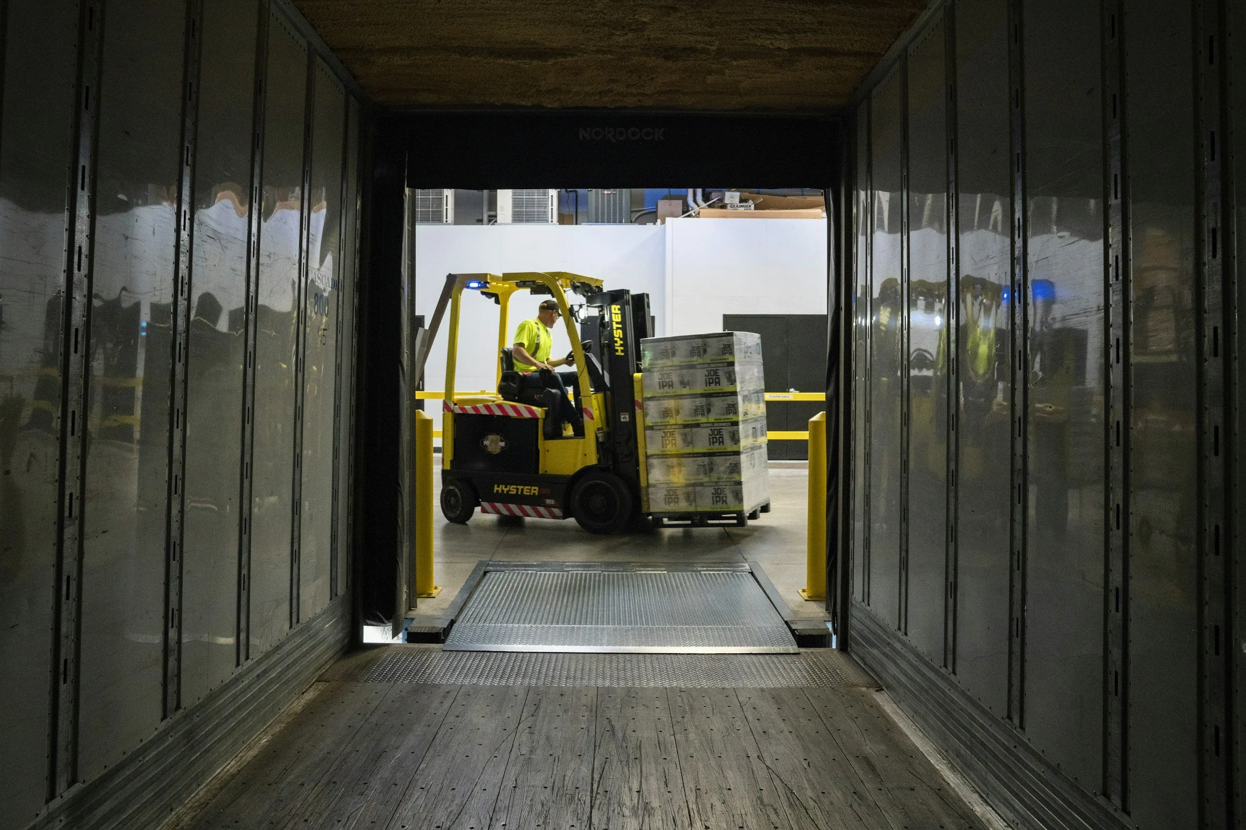 View through a dark corridor to a warehouse or loading bay where a person is operating a yellow forklift carrying boxes, with reflective metal walls on each side.