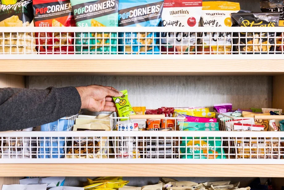 A person reaching for a green gummy candy package on a snack shelf in a store, with various snack bags organized on multiple white wire shelves.
