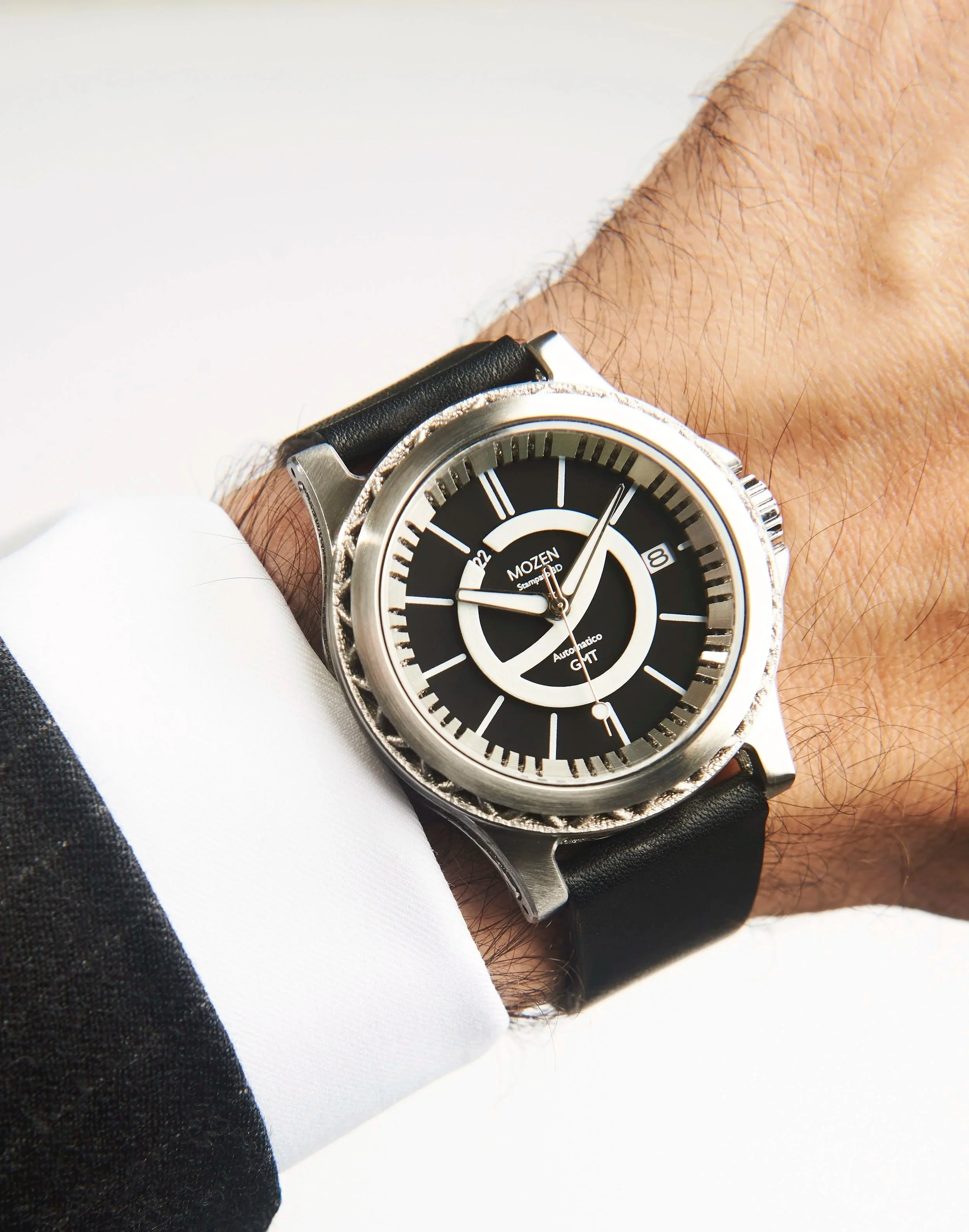 A close-up of a wristwatch with a black dial, silver case, black strap, and multiple time zones, worn on a person's wrist dressed in a black suit and white shirt.
