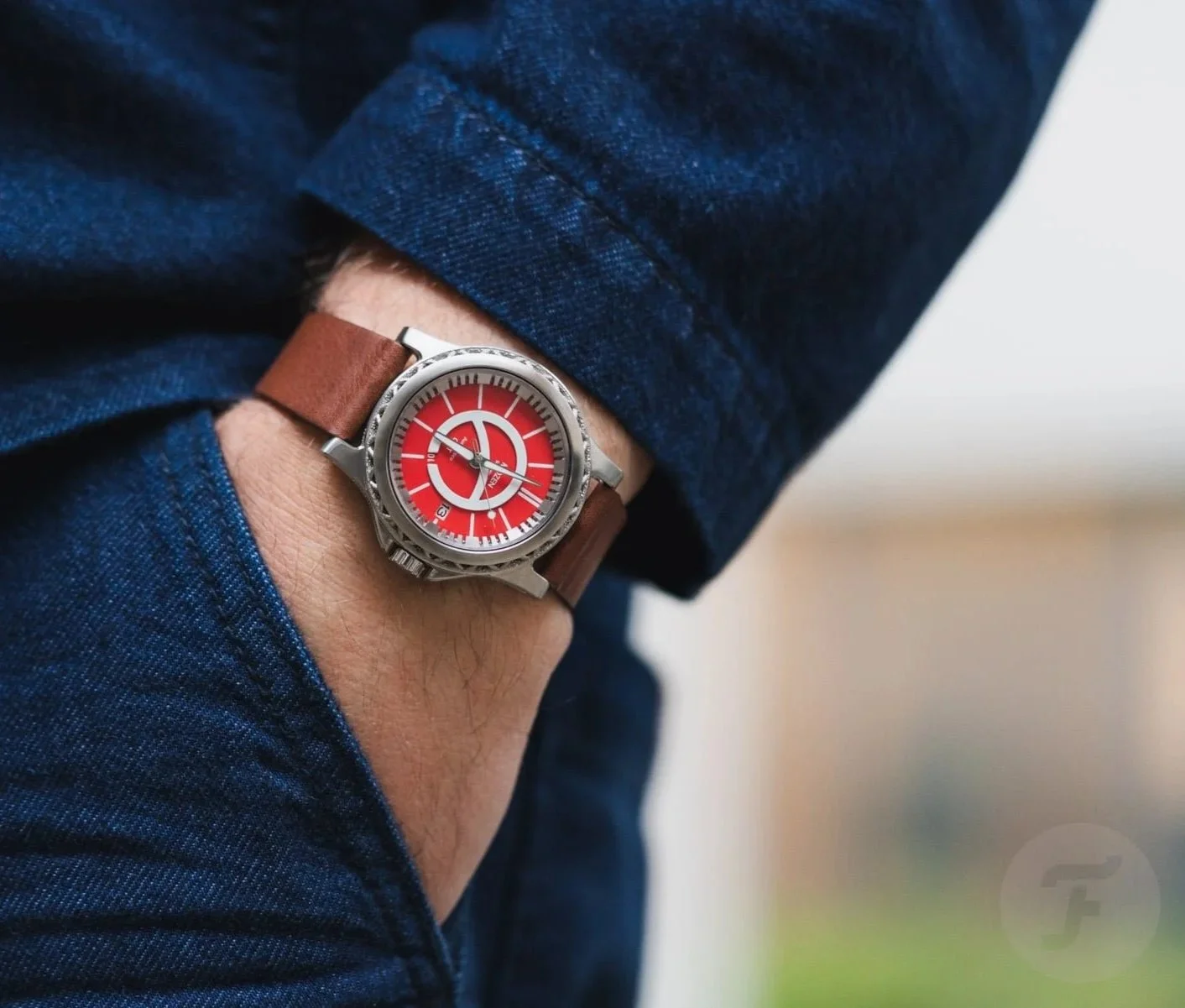A person's wrist wearing a watch with a brown leather strap and a red dial, partly covered by a blue denim shirt sleeve.