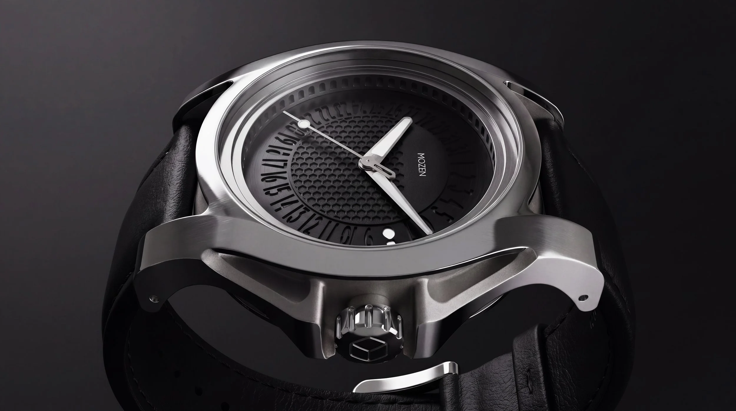 Close-up of a modern wristwatch with a black dial, silver case, and black leather strap. The brand 'MOZEN' is visible on the dial. The watch has a unique design with a rotating date disc and textured center.