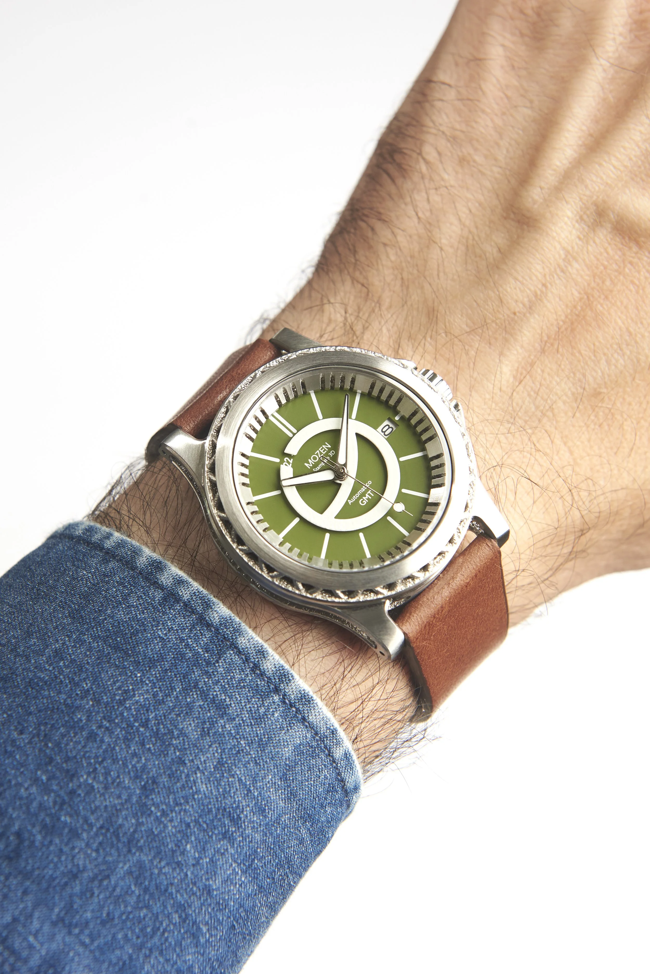 A wristwatch with a green dial, silver bezel, and brown leather strap worn on a man's wrist with visible arm hair, wearing a blue denim sleeve.