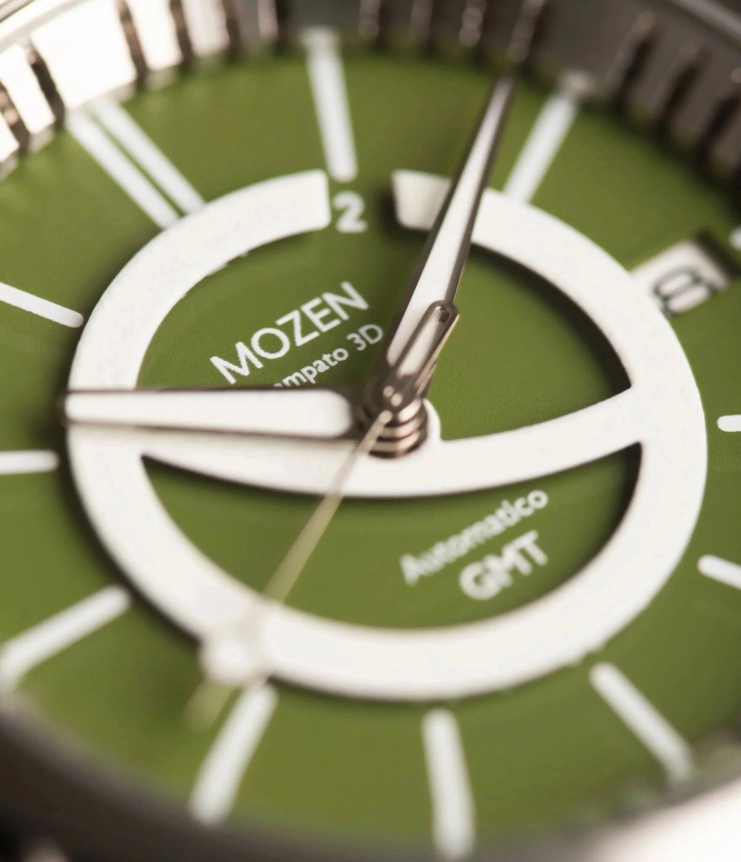 Close-up of a green Mozen watch face with white hour markers and hands, displaying the time at approximately 11:58, with text indicating 'Mozen' and 'comparto 3D'.