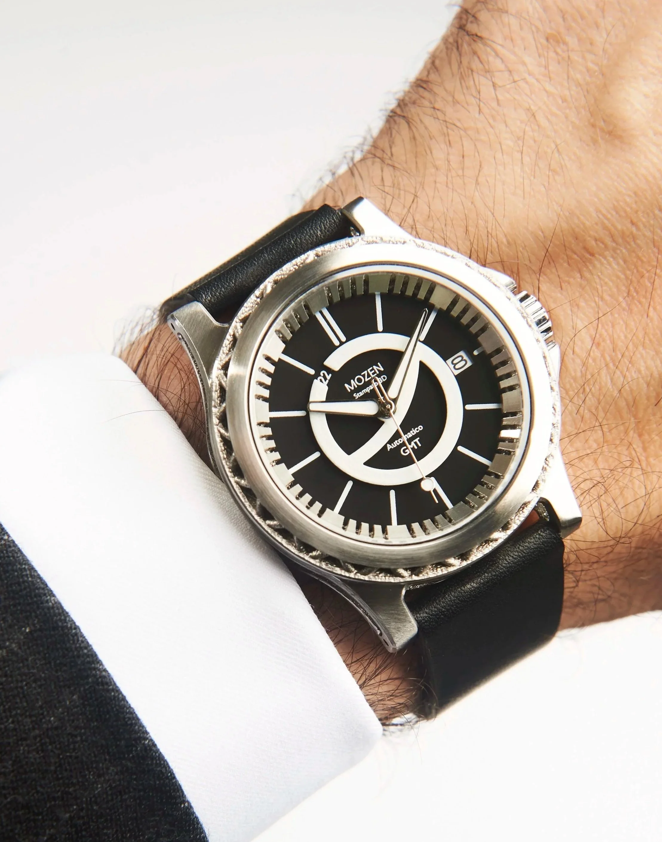 A wristwatch with a black dial, silver case, and black leather strap worn over a white shirt cuff and black suit sleeve.