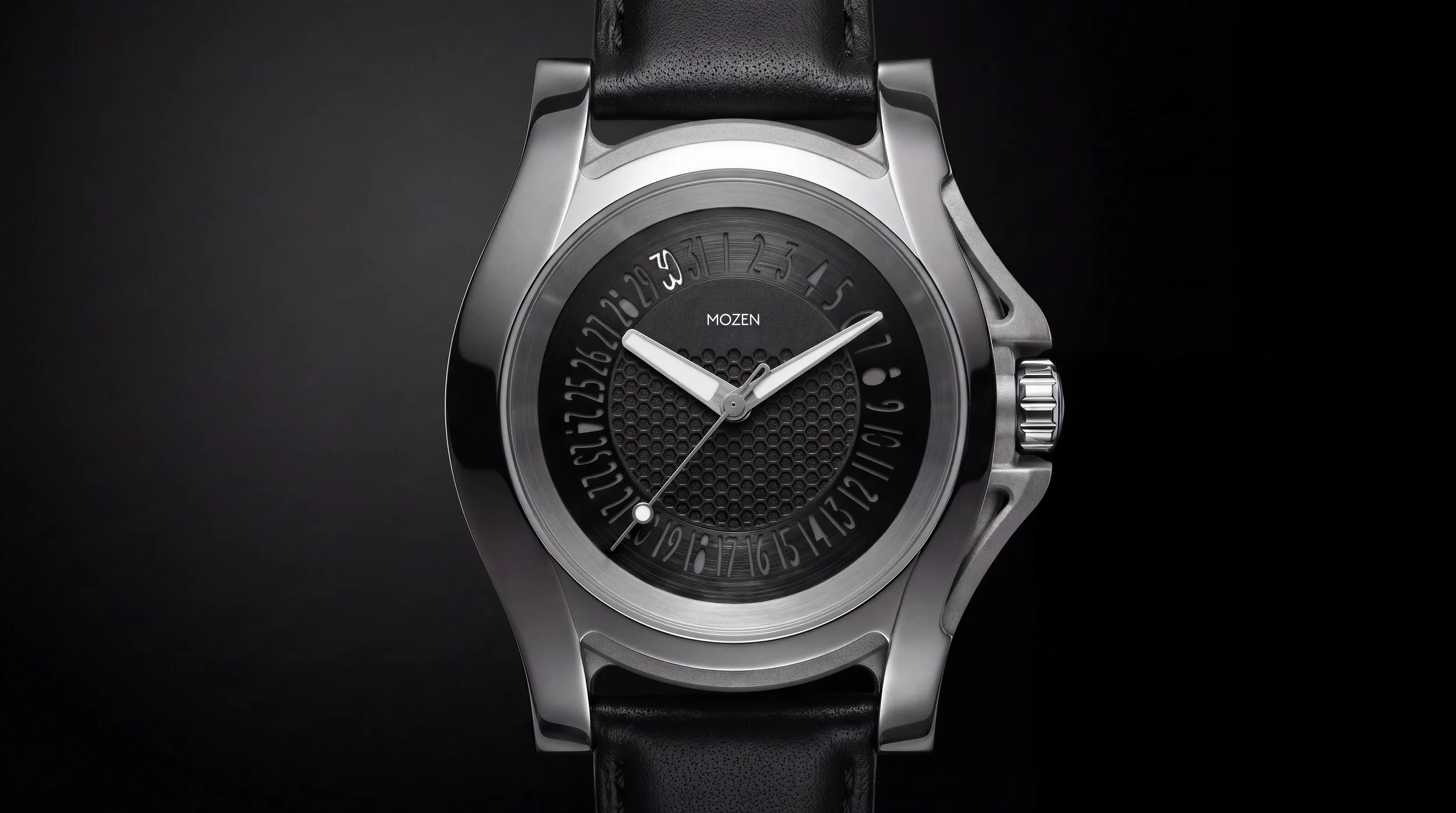 A modern wristwatch with a black dial and silver case, featuring a textured pattern and unique dial with numbered date display, on a black strap against a dark background.