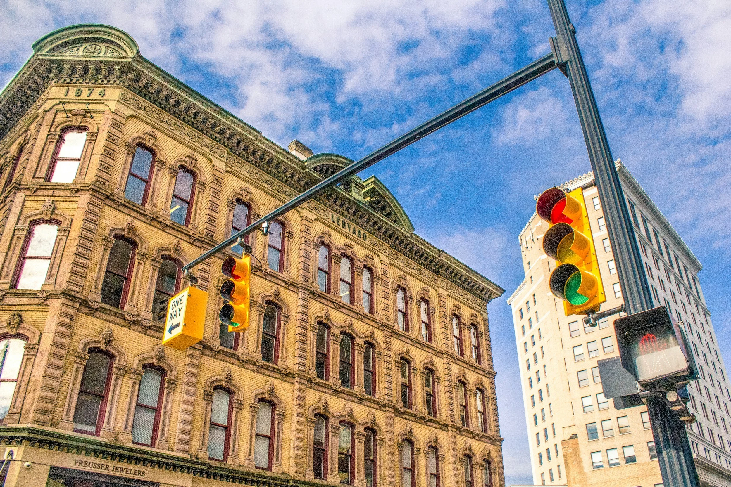 City street scene with historic brick building, modern high-rise, traffic lights, and a one-way street sign against a partly cloudy blue sky.