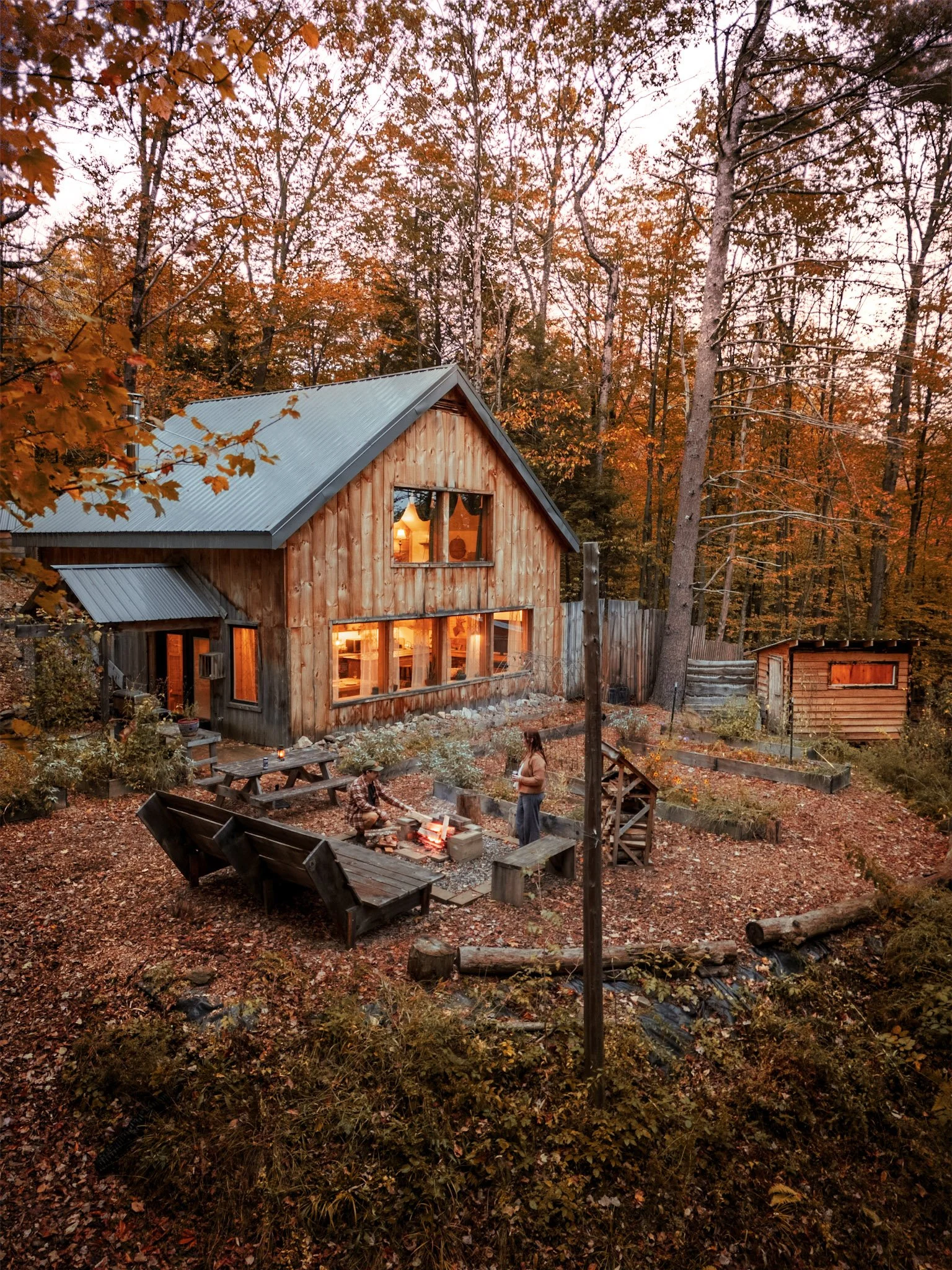 A wooden cabin with large windows, surrounded by autumn trees with orange leaves, with people sitting and standing by a fire pit outside.
