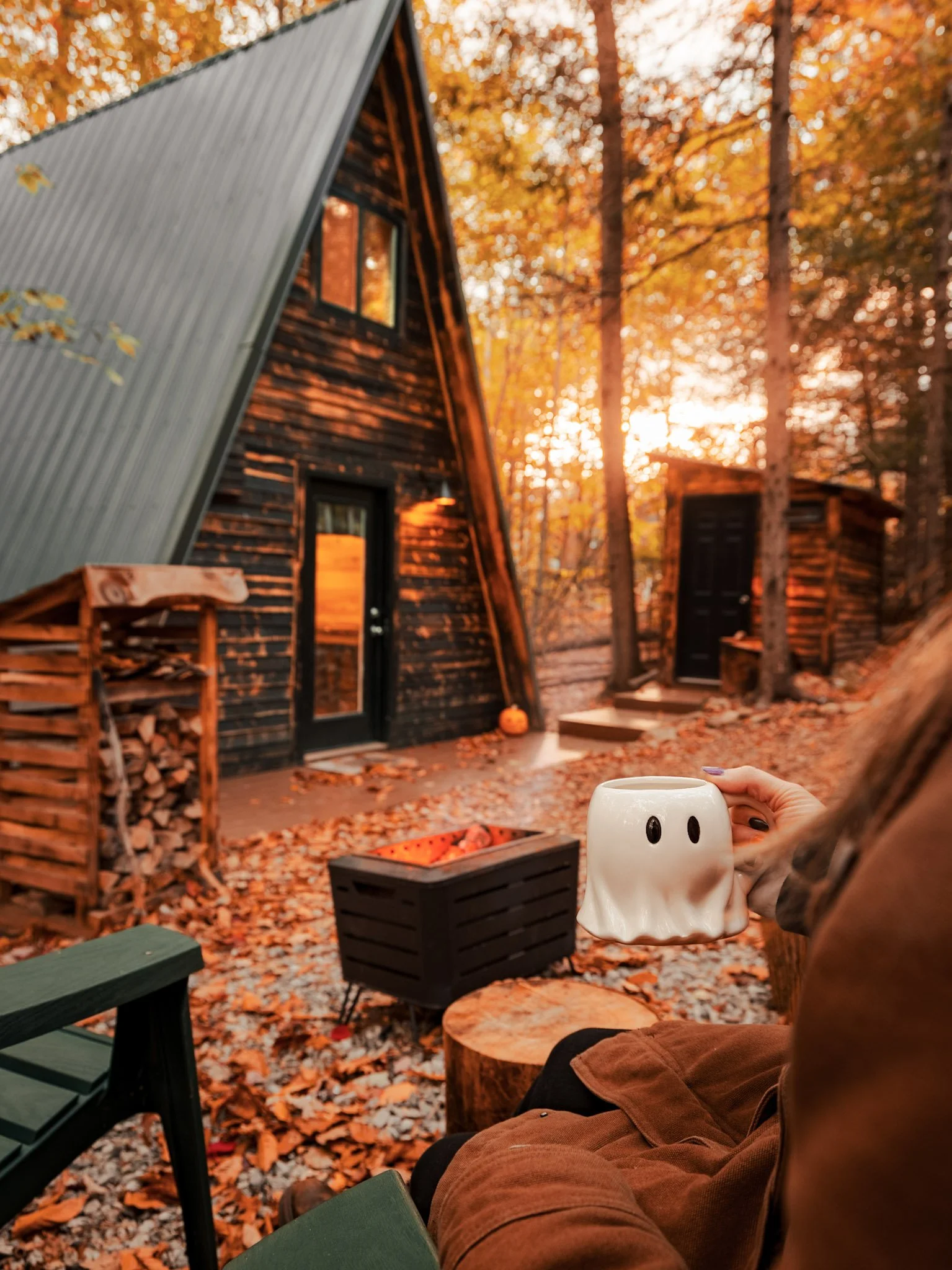 A person holding a ghost-shaped mug outside a rustic A-frame cabin in a wooded area with fall leaves on the ground and trees in the background in autumn.