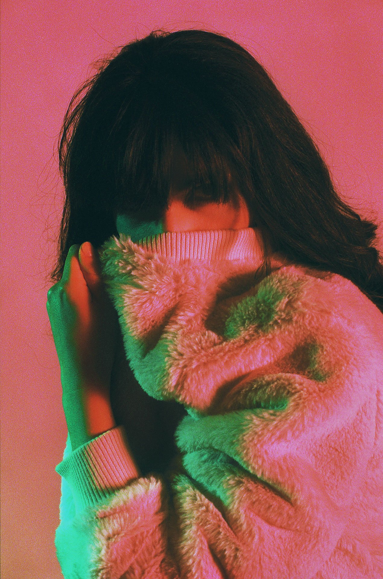 A woman with dark hair covering her face, wearing a cozy, fluffy pink sweater, holding a finger to her lips, against a pink background with green lighting.