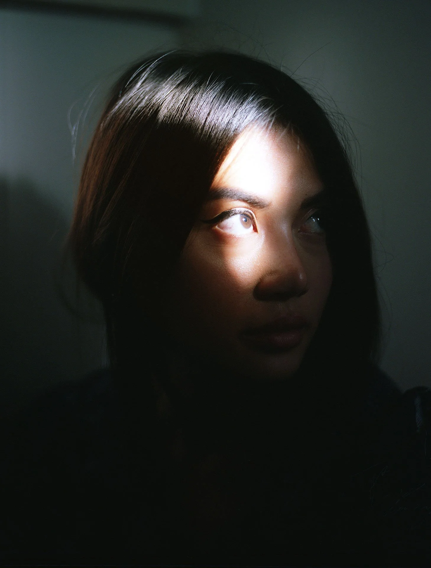 A woman with dark hair and light-colored eyes looking upwards with sunlight hitting her face.